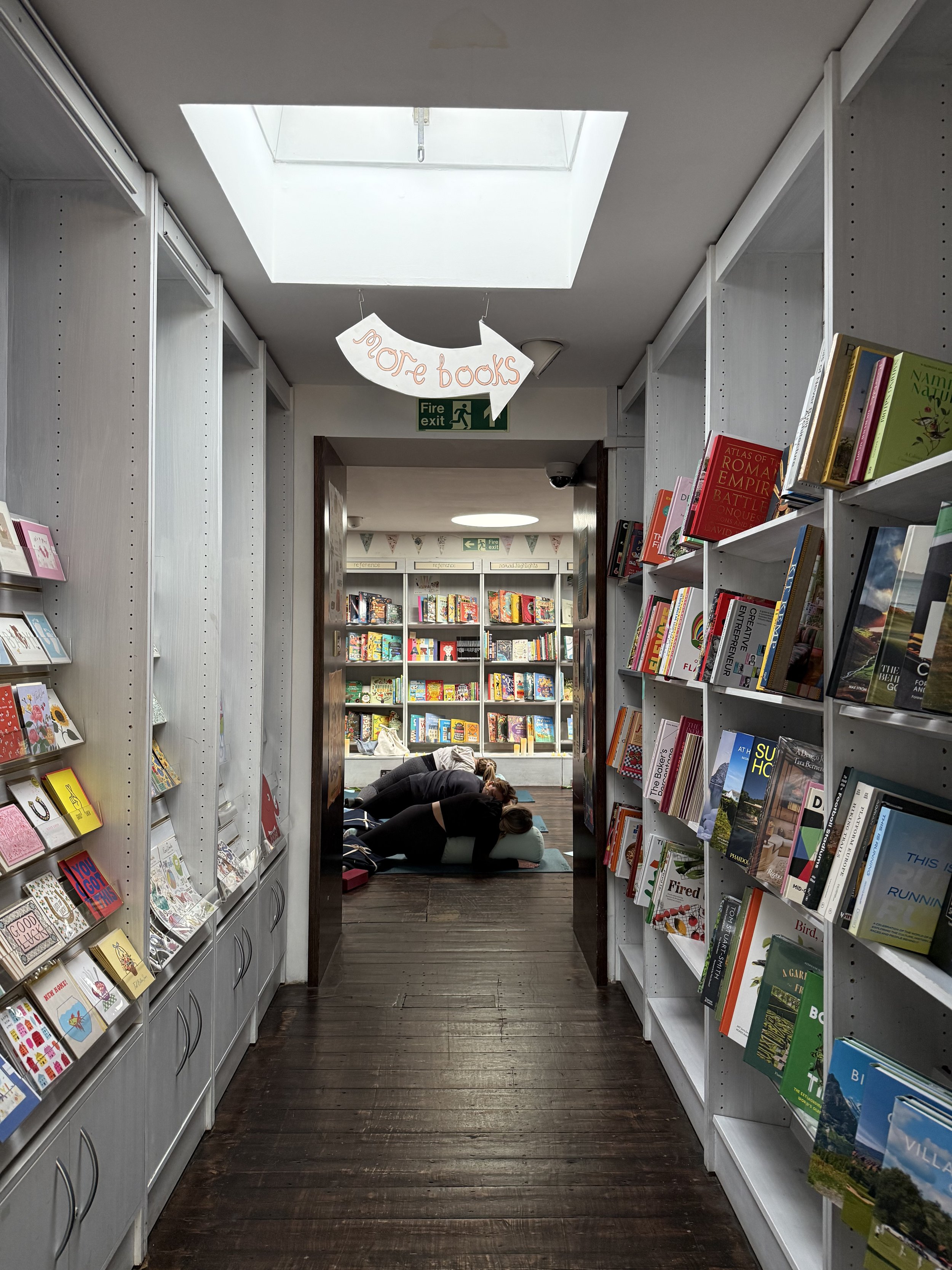 Bookshelf aisle in a bookstore with people practicing yoga on mats in the background. Sign pointing to 'more books' hanging from ceiling.