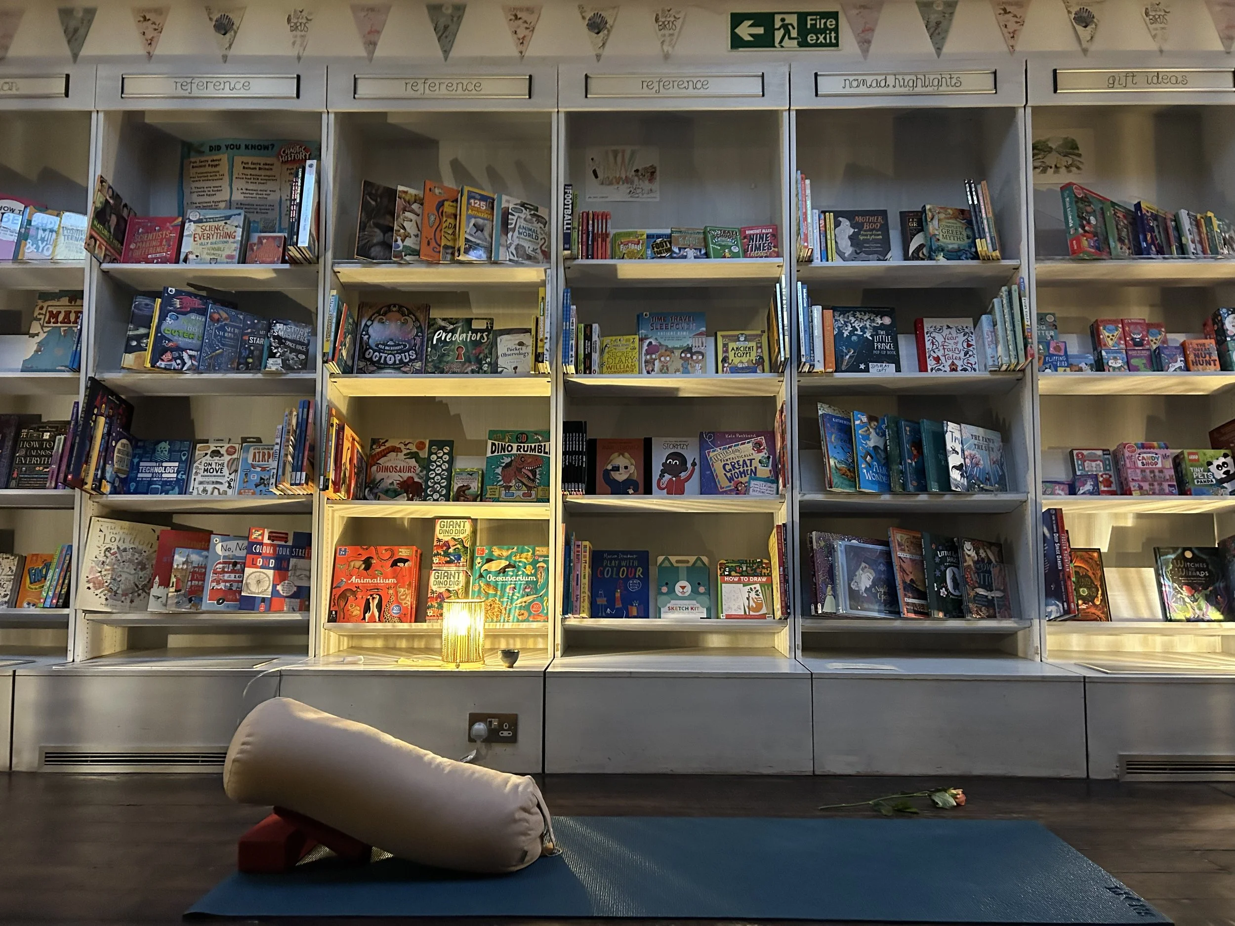 Children’s bookstore or library with shelves of colorful books, a reading area with blue mats and cushions, triangular bunting, and a fire exit sign.