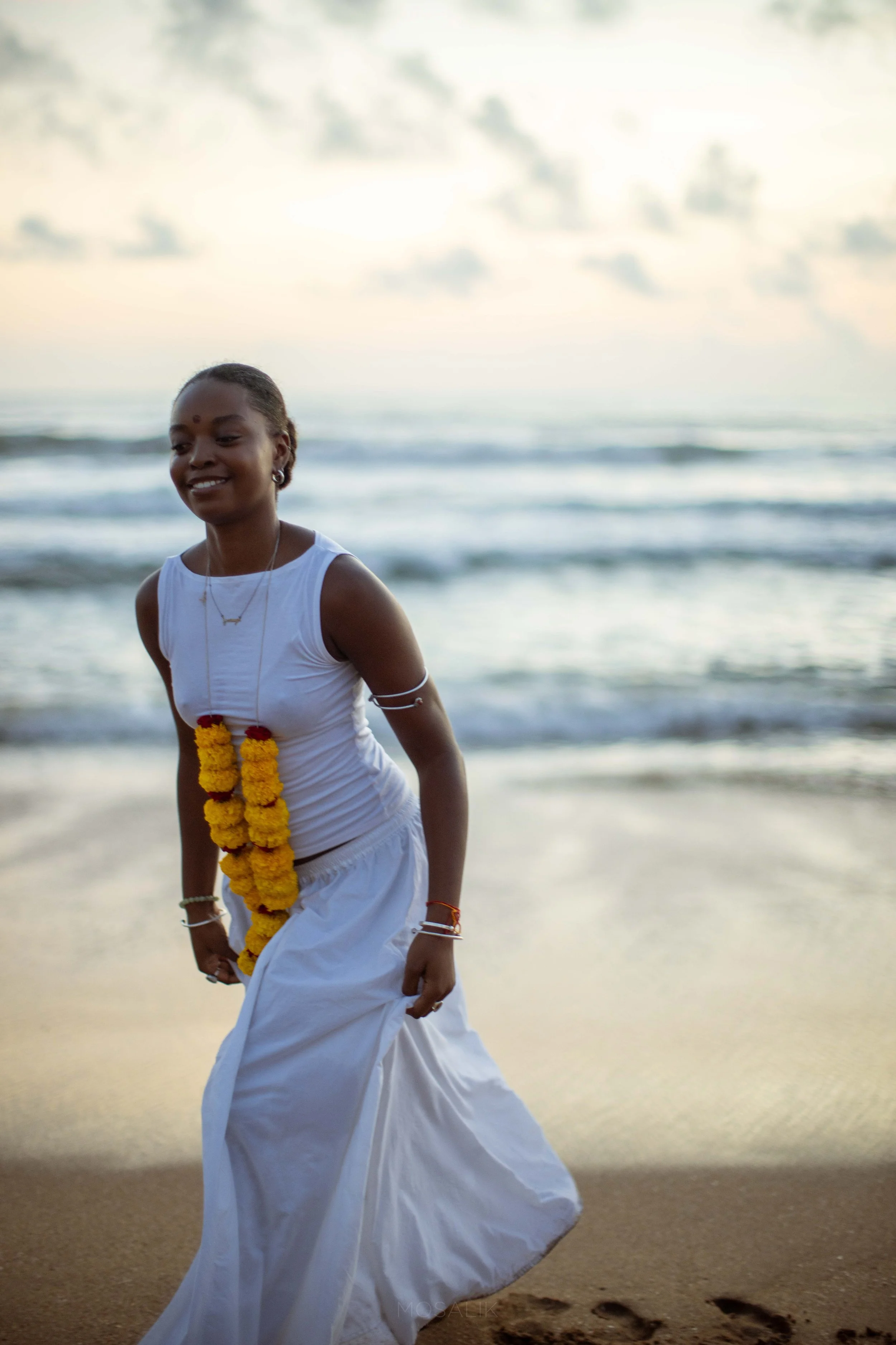 A woman dressed in traditional Indian attire, walking along the beach at sunset, wearing white clothing and yellow floral garland.