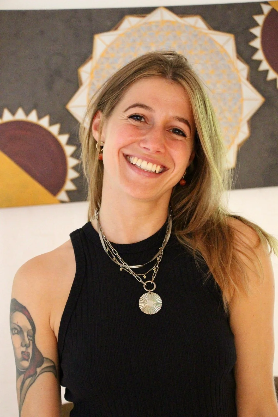 A smiling woman with long light brown hair, wearing a black sleeveless top and layered silver necklaces, standing in front of an abstract geometric art piece.