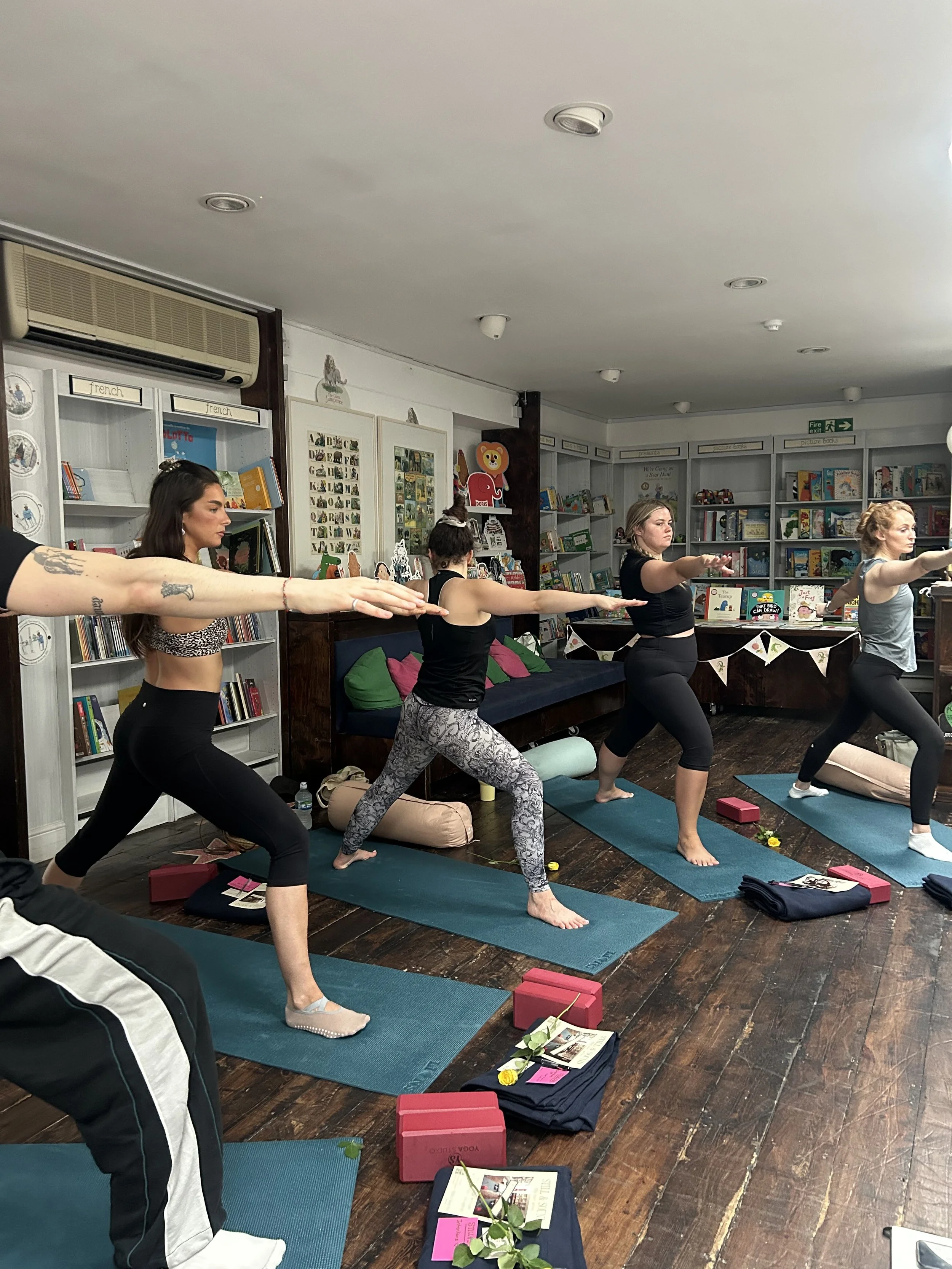 Group yoga class practicing lunges on mats inside a children's bookstore surrounded by bookshelves and yoga blocks