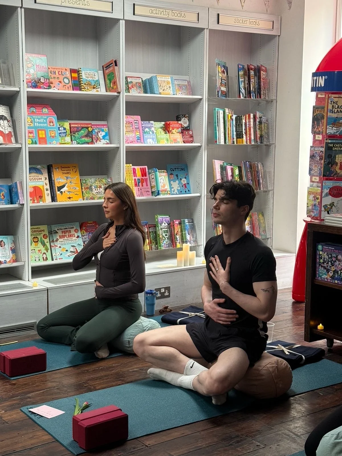 Two people sitting on yoga mats in a library-like space, meditating with their hands on their chests. Bookshelves with children's books are in the background, and candles are placed behind them.