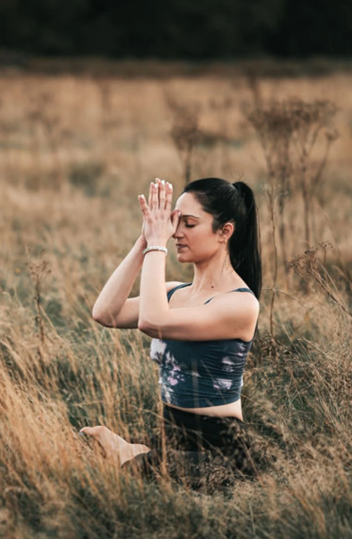 A woman practicing yoga outdoors in a field with tall grass, sitting with her eyes closed, hands pressed together in prayer position against her forehead.