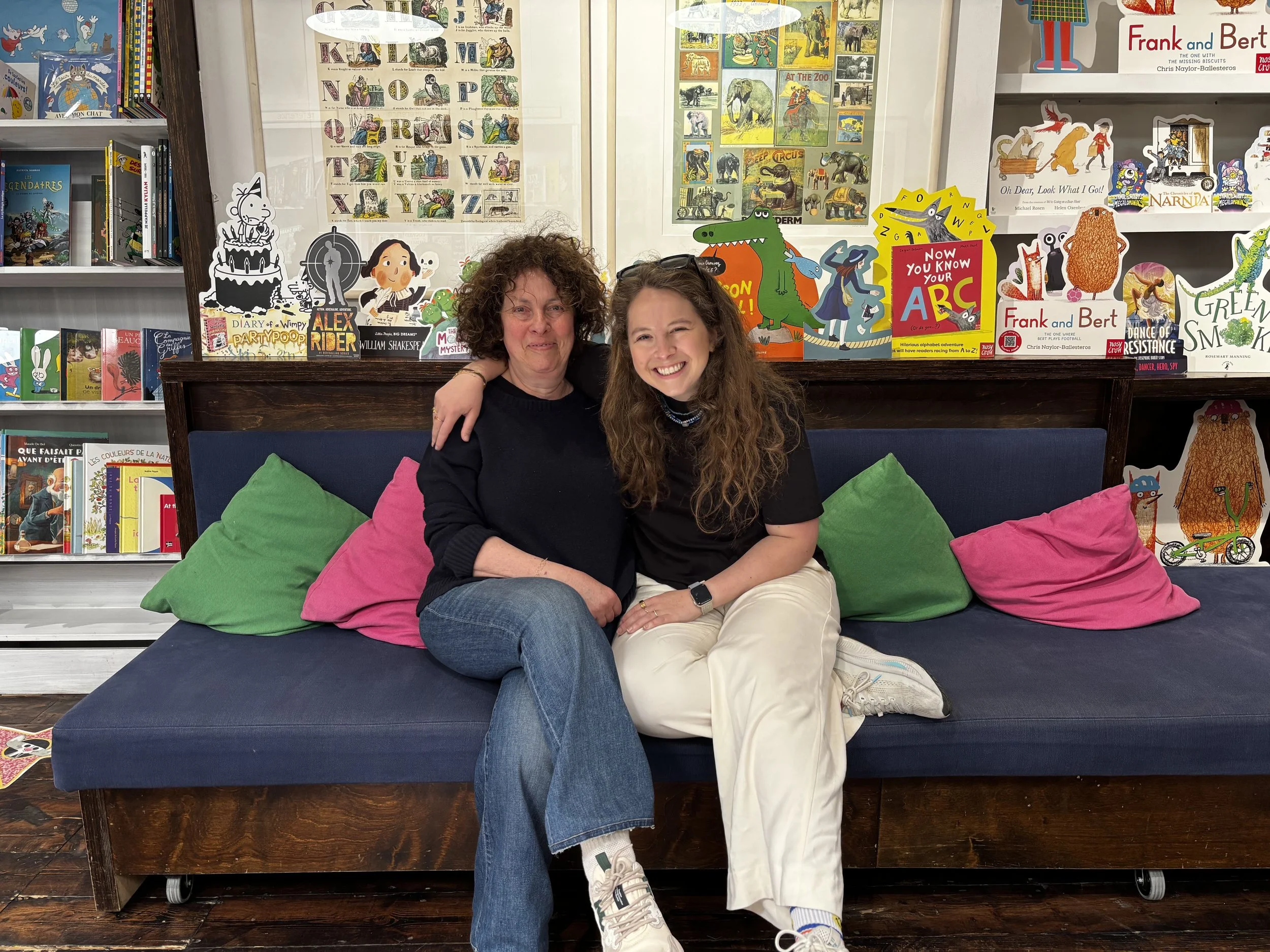 Two women sitting on a dark blue bench with colorful pillows, smiling, in front of a glass display case filled with children's books and colorful illustrated posters featuring animals and alphabet. The shelves behind contain more books and decorations related to children's literature.