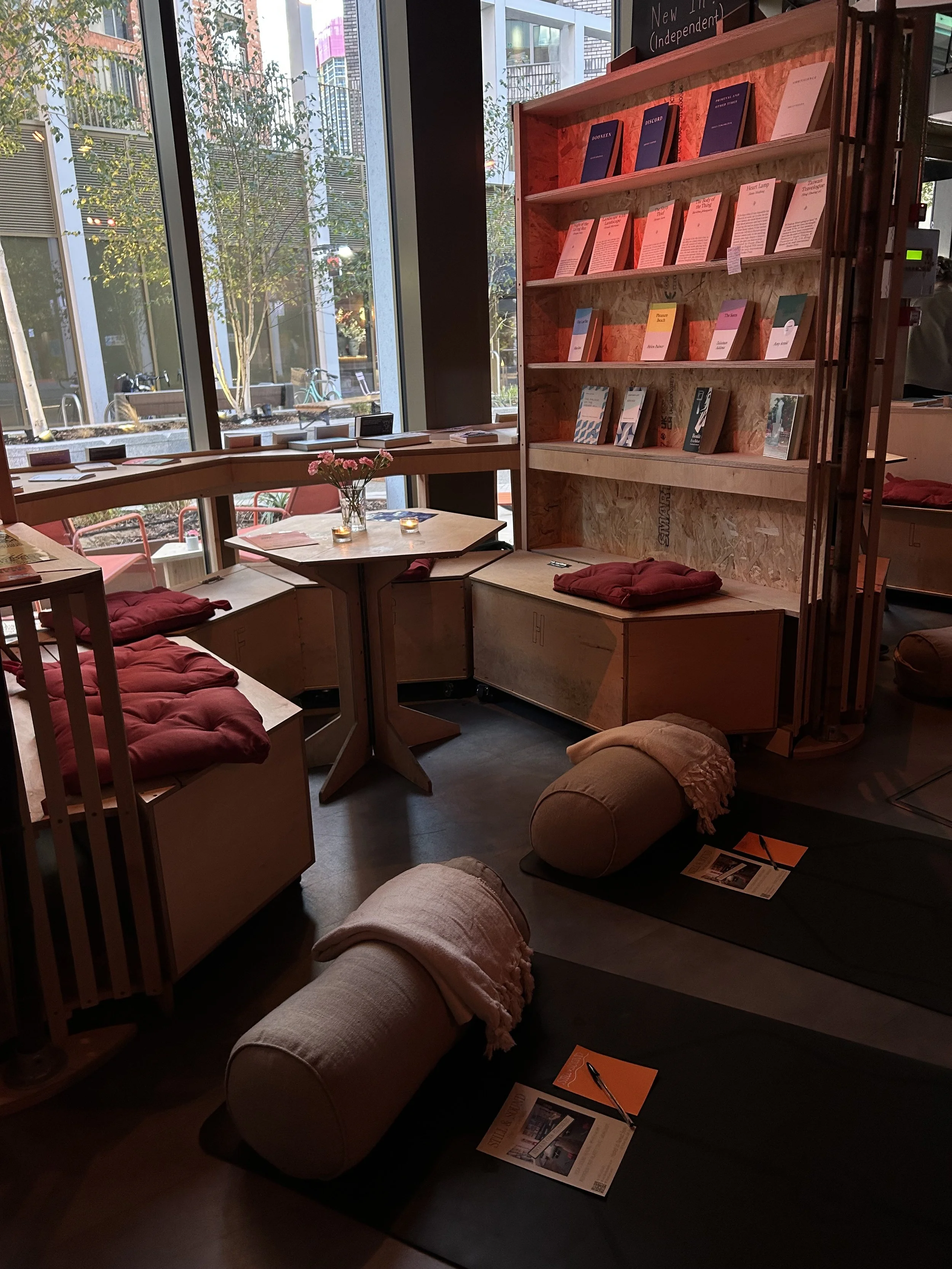 Cozy independent bookstore reading lounge with bookshelves, cushioned wooden benches, tables, candles, and large windows overlooking an urban courtyard