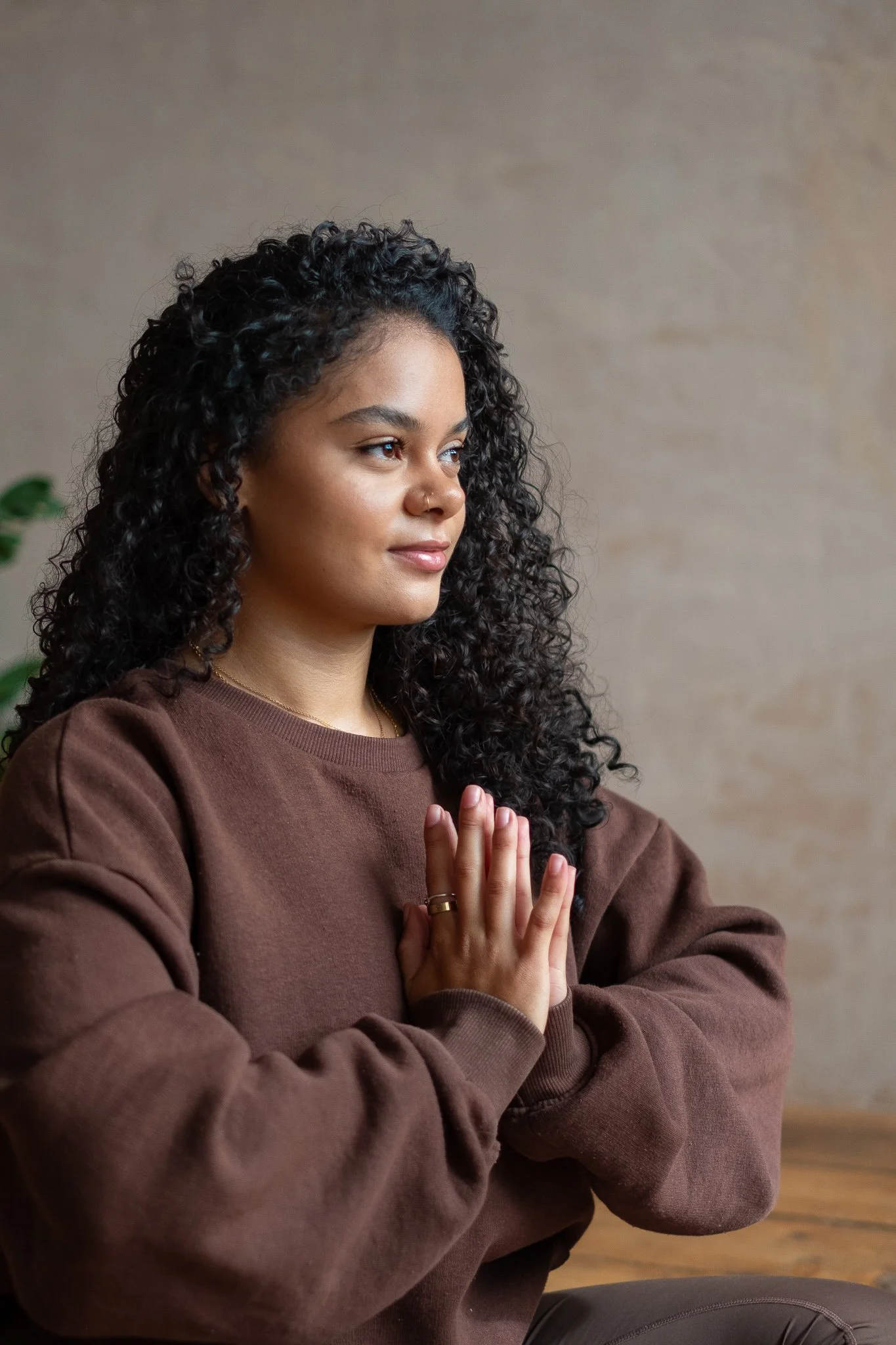 A woman with curly dark hair and a nose piercing is sitting with her eyes slightly closed, hands pressed together in a prayer or meditation position. She is wearing a brown sweatshirt and has a calm, focused expression. The background features a neutral wall and a hint of green plant to the left.