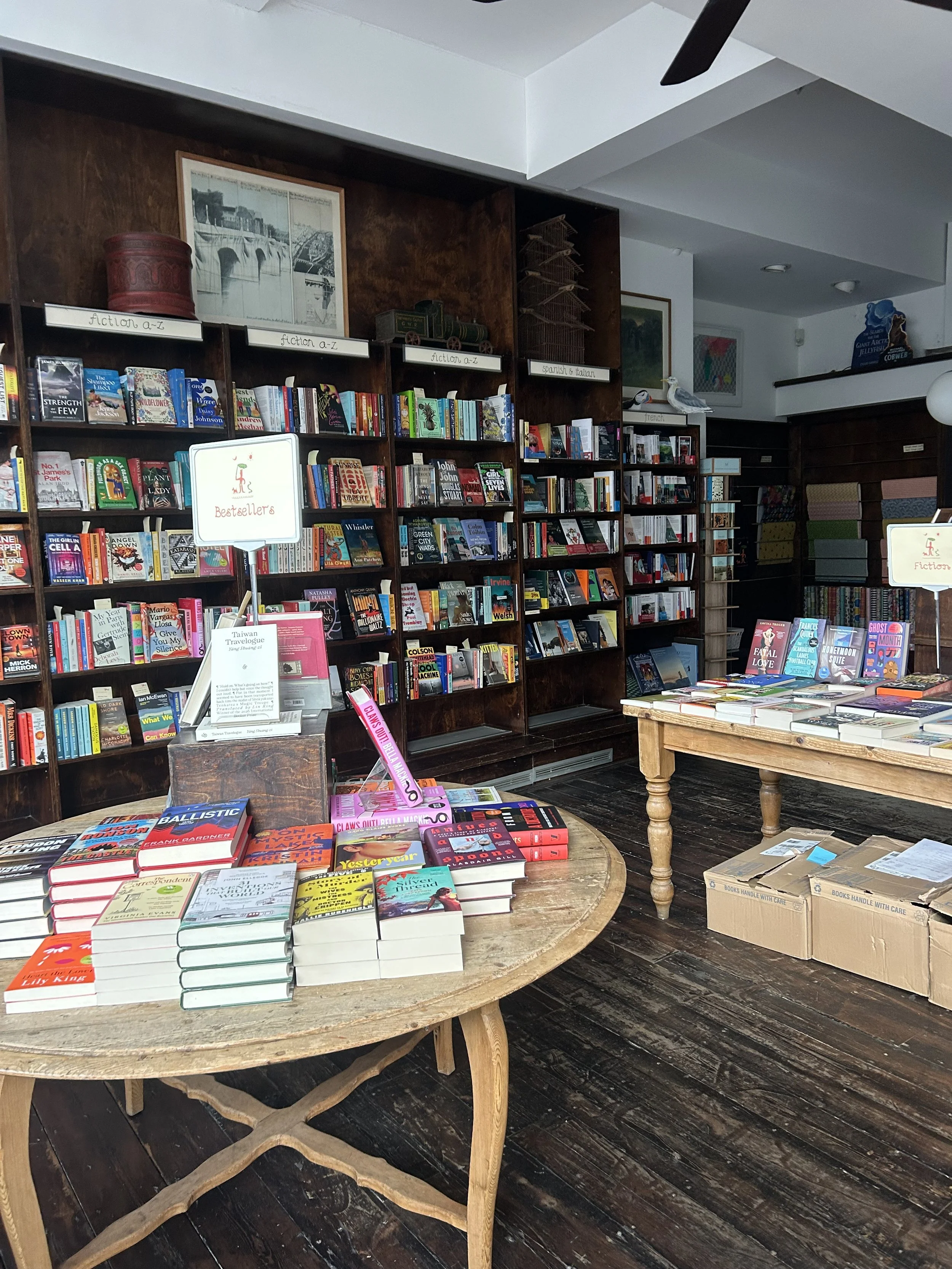 Interior of an independent bookstore with floor-to-ceiling bookshelves, fiction and foreign-language sections, and tables displaying new books.