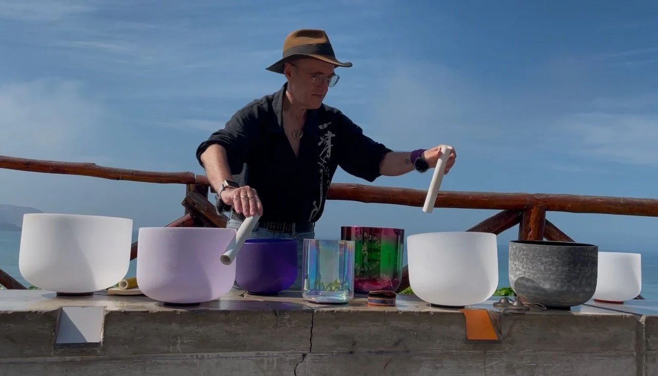 A person wearing a hat and glasses is playing singing bowls on a wooden outdoor table with a scenic ocean view in the background.