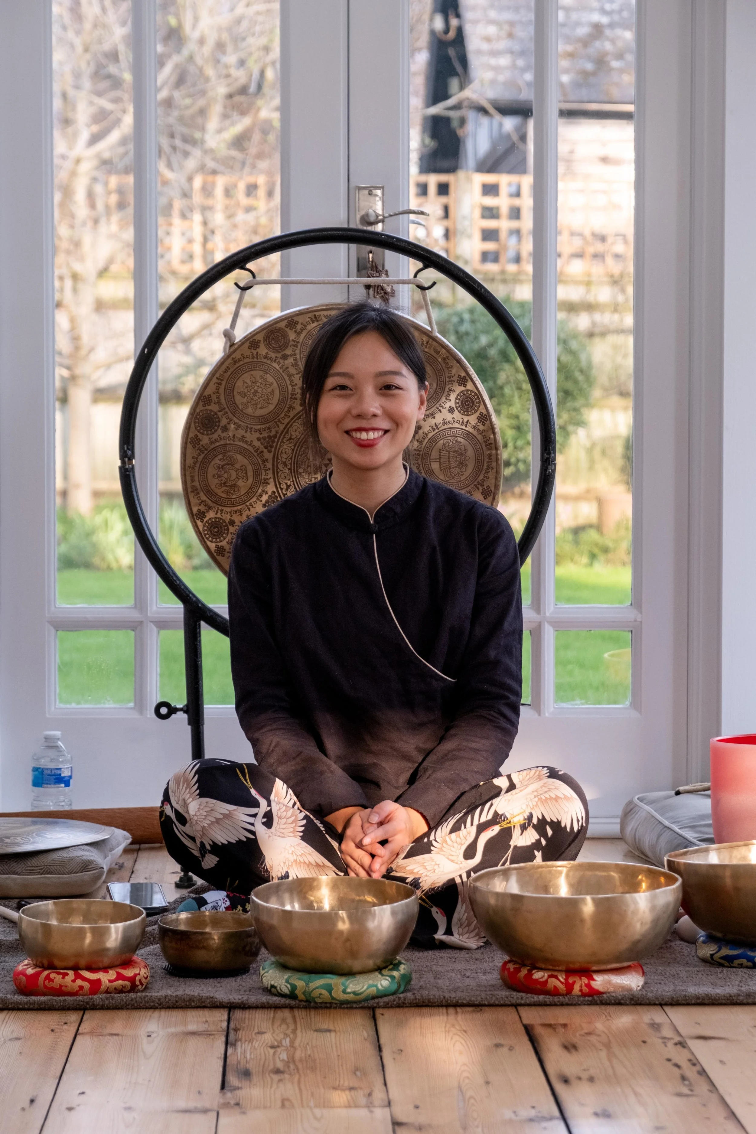 A woman smiling and sitting cross-legged on the floor in front of a window with a garden view. She is surrounded by singing bowls placed on colorful cushions, with some objects and a water bottle nearby.