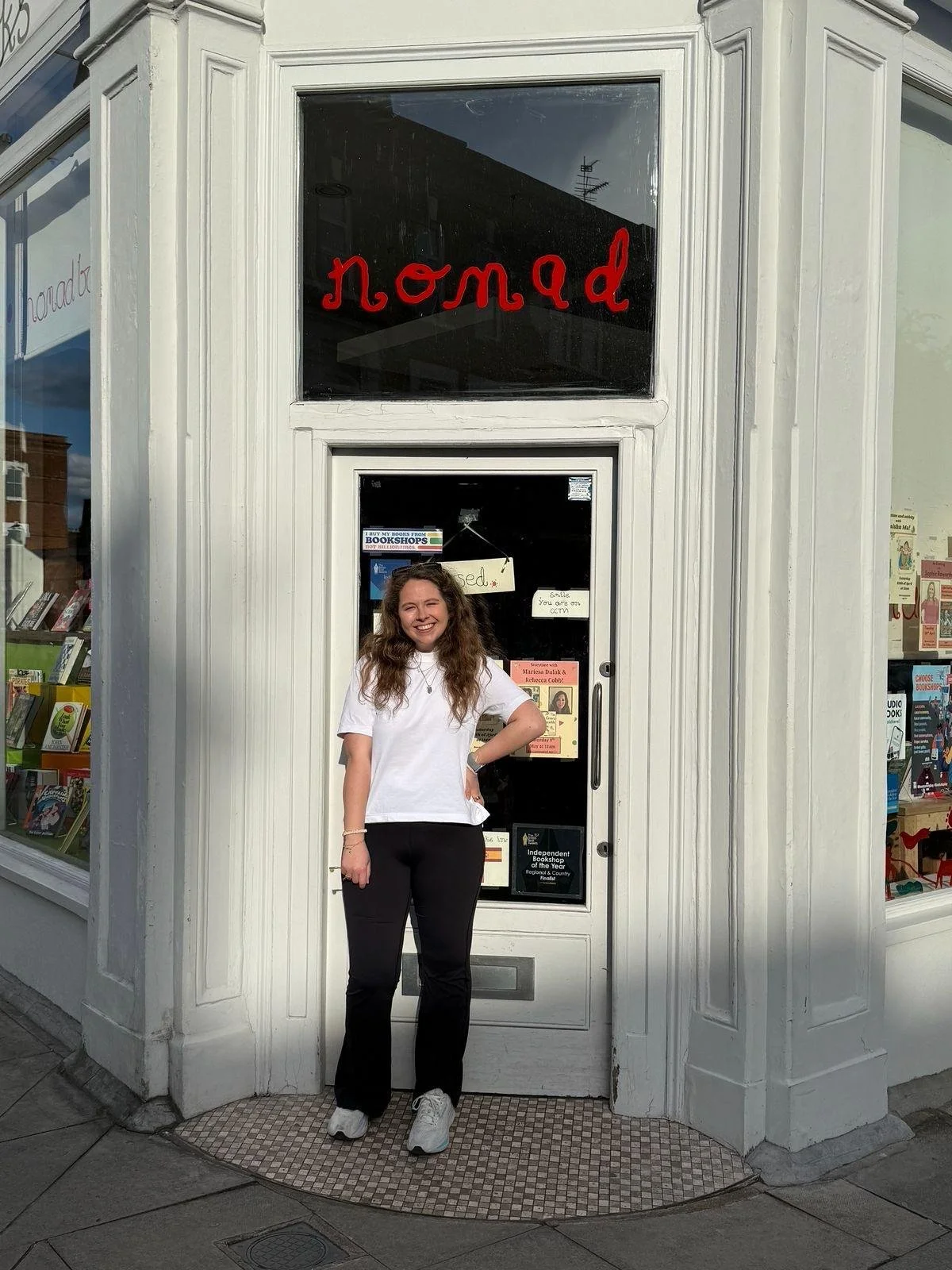 A smiling woman with long curly hair wearing a white t-shirt and black pants standing in front of a white bookstore storefront with the word 'nomad' written in red above the entrance. The bookstore window displays books and signs.