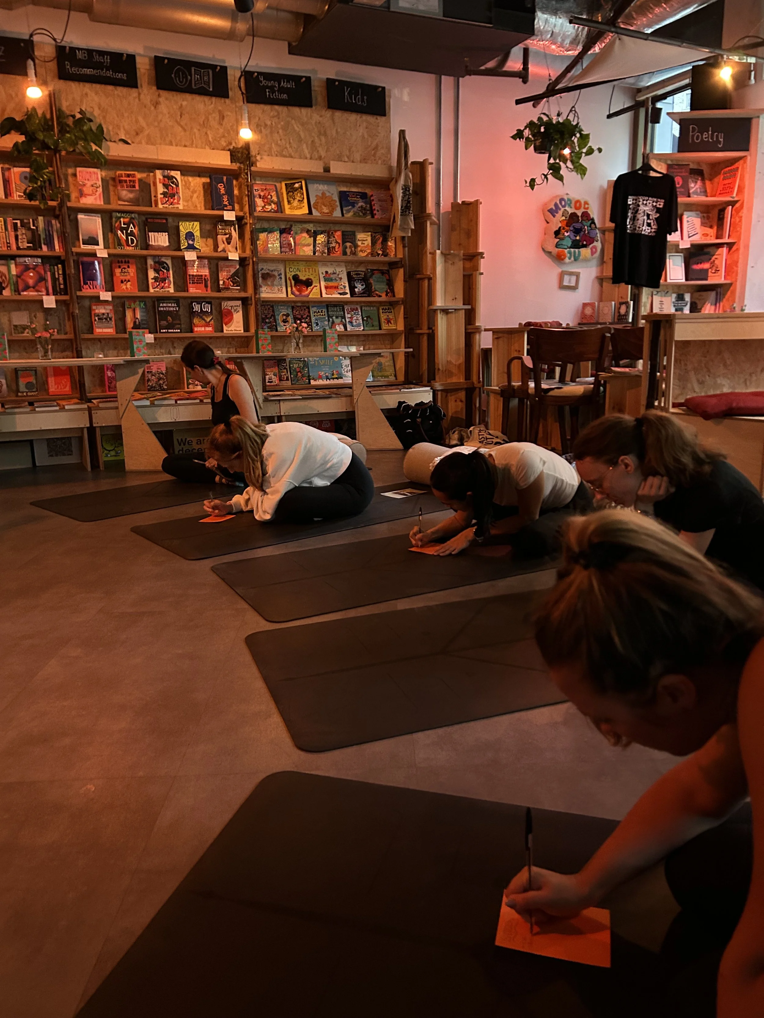 People participate in a writing workshop inside an independent bookstore, surrounded by bookshelves, category signs, plants, and warm hanging lights.