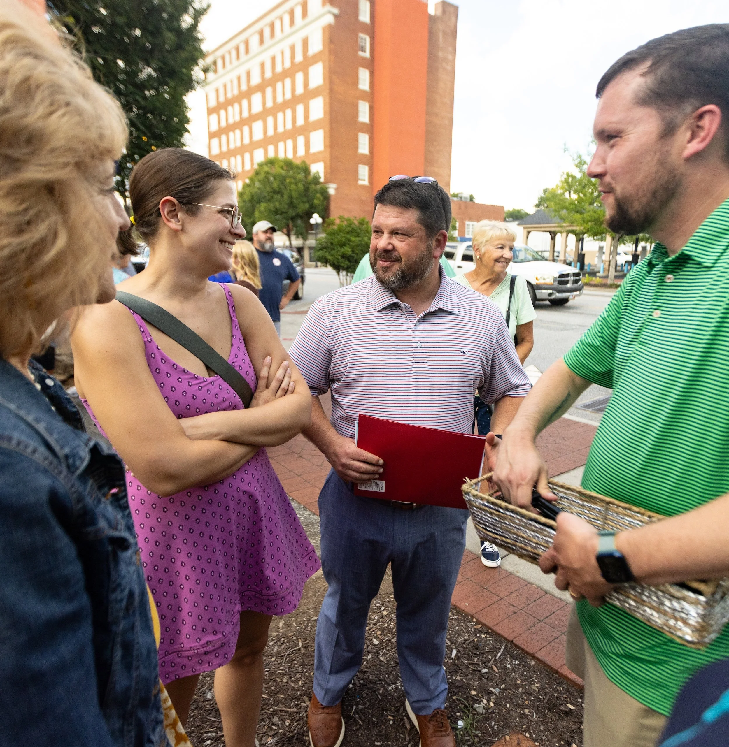 Group of people talking outdoors on city street with tall brick building and trees in background.