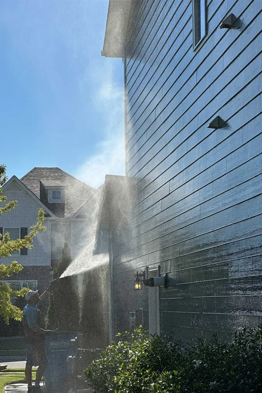 A person using a pressure washer on the side of a house, spraying water onto the exterior wall with a blue sky and neighboring houses in the background.