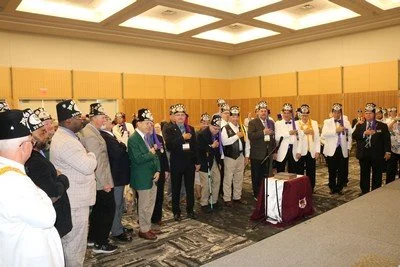 A group of people wearing crowns and formal attire participating in a ceremony or celebration in a large, well-lit room.