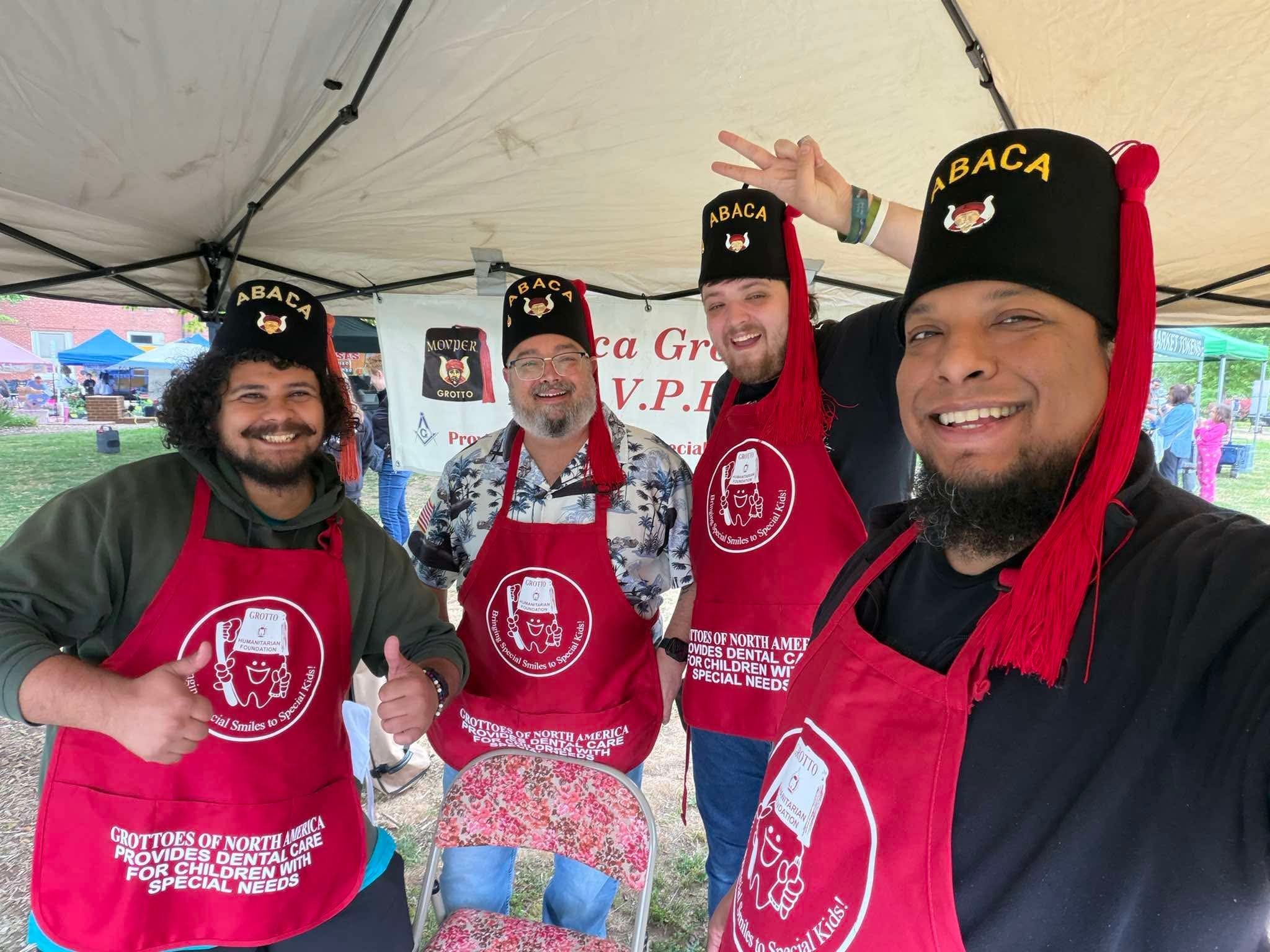 Four smiling men wearing red aprons and black hats with 'ABACA' on them, standing under a canopy at an outdoor event, with tents and people in the background.