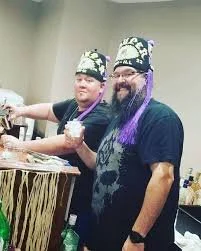 Two men with beards and head coverings posing together in a room, one holding a small glass container.