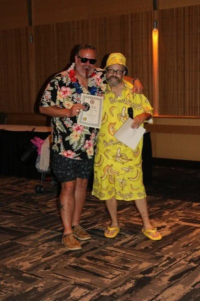 Two men dressed in colorful, tropical-themed outfits standing together indoors. One wears a floral shirt, sunglasses, and holding a certificate, while the other wears a banana-patterned dress, headscarf, and glasses, holding a piece of paper.