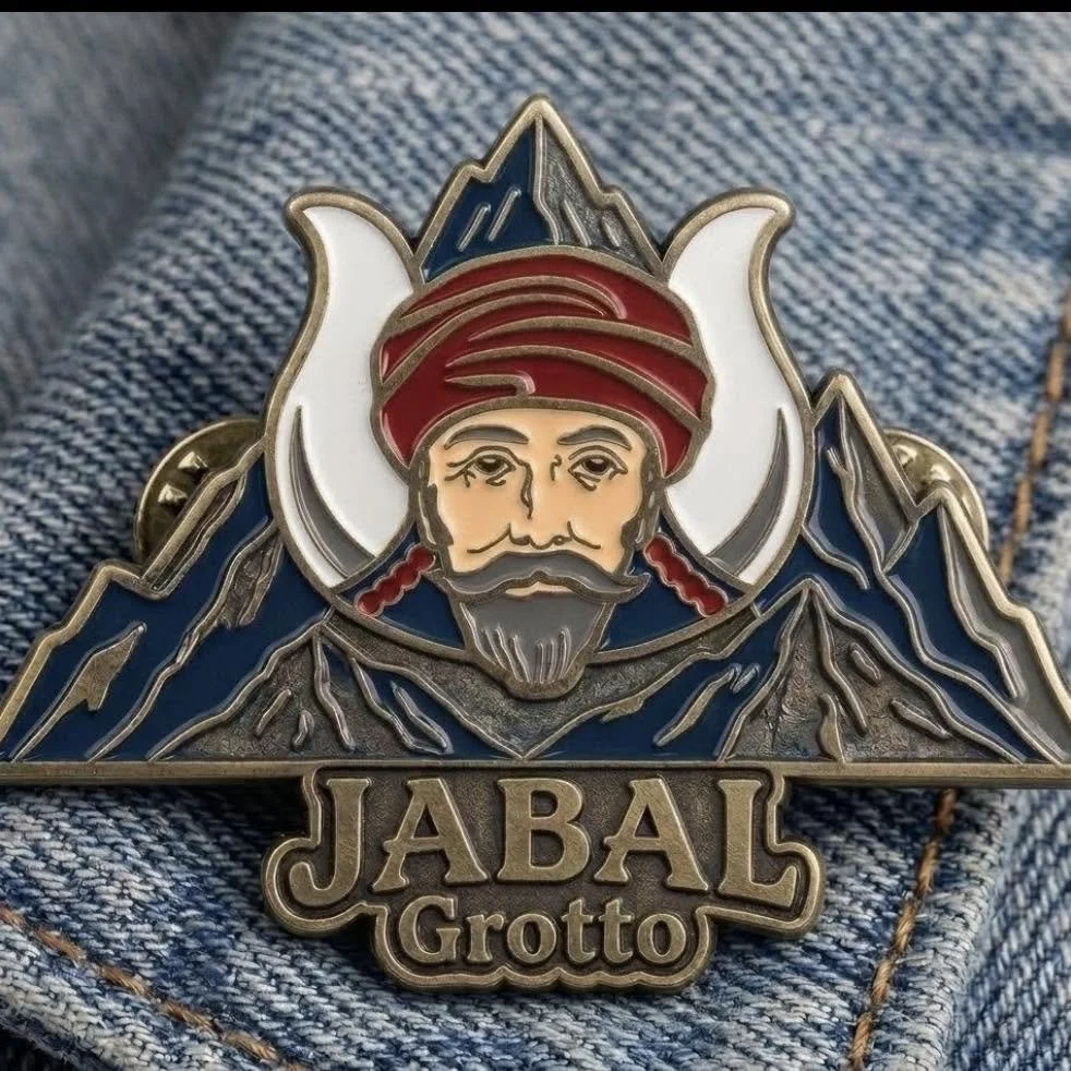 A metallic badge featuring a mountain landscape with a man's face wearing a red head wrap, and the words 'JABAL Grotto' below.