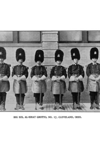 Historical black and white photograph of six soldiers in uniform standing in a line outside a building in Cleveland, Ohio, with a caption describing them as Big Six Al-Niqat Grotto members.
