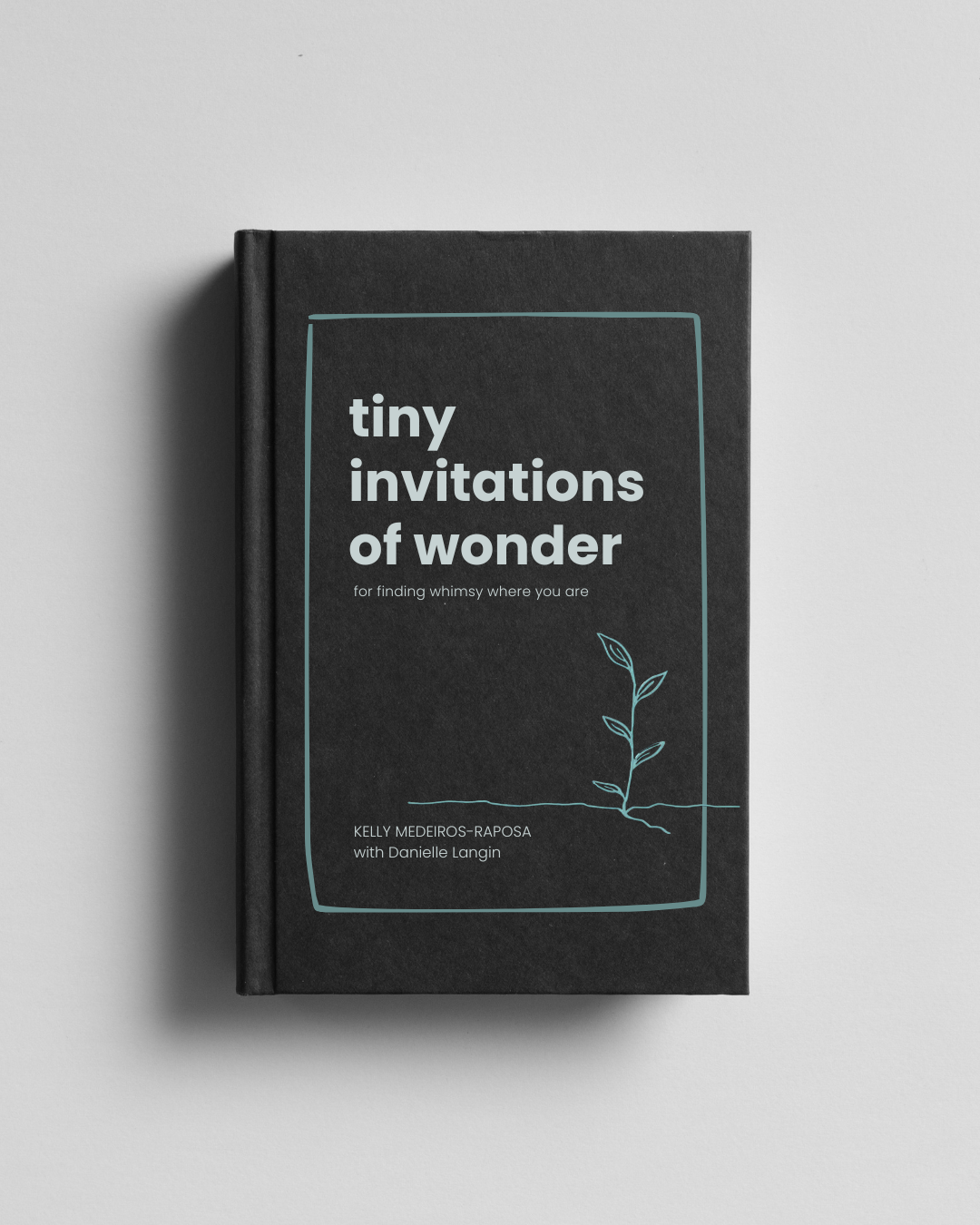 A black hardcover book titled "tiny invitations of wonder" authored by Kelly Medeiros-Raposa with Danielle Langin with a minimalist cover design featuring a small plant illustration, placed on a white surface.