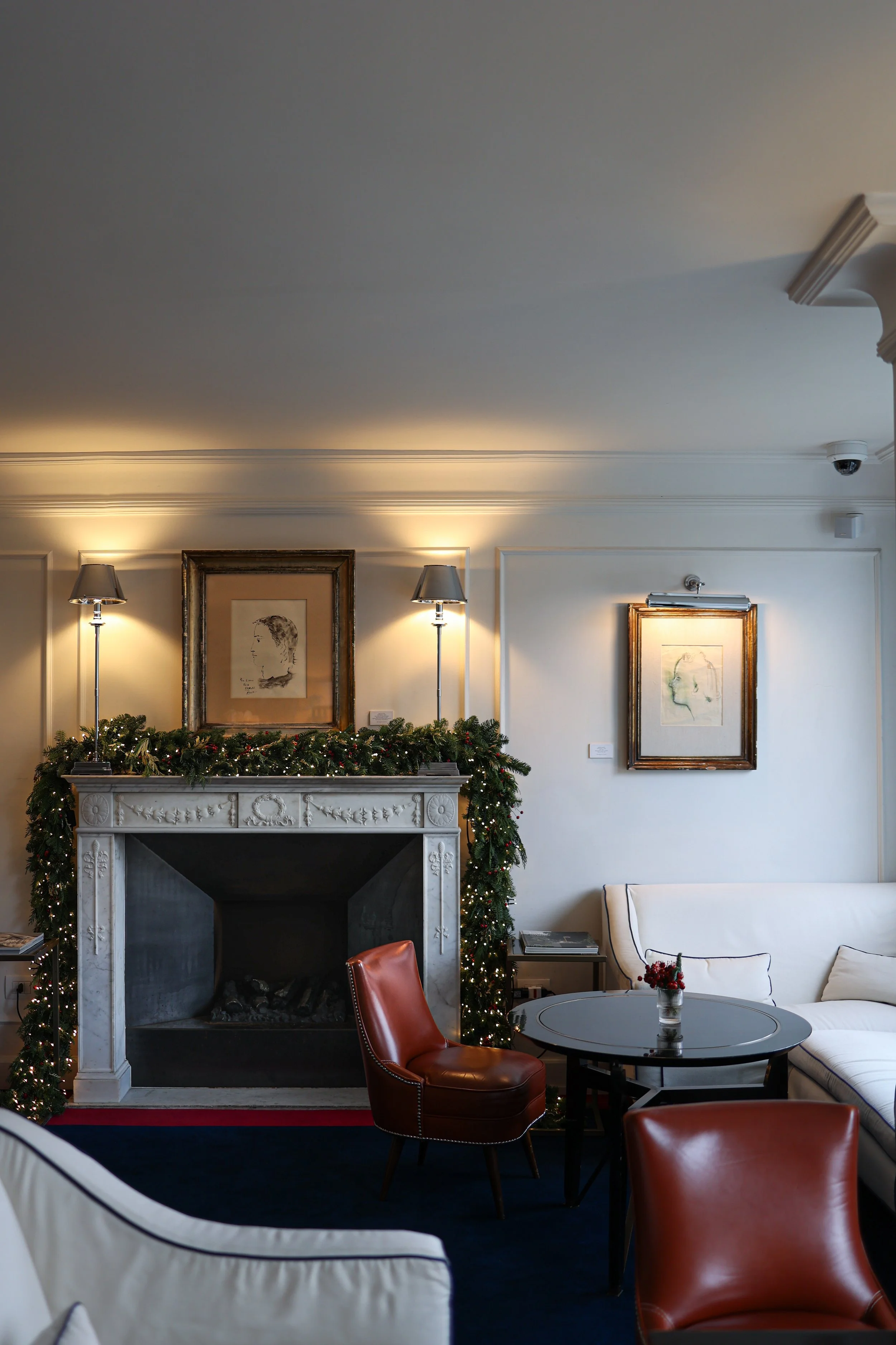 Interior of a room with a decorated fireplace, framed artwork on the wall, a white sofa, a round coffee table with a small flower arrangement, and red chairs. Holiday garland and string lights adorn the fireplace mantel.