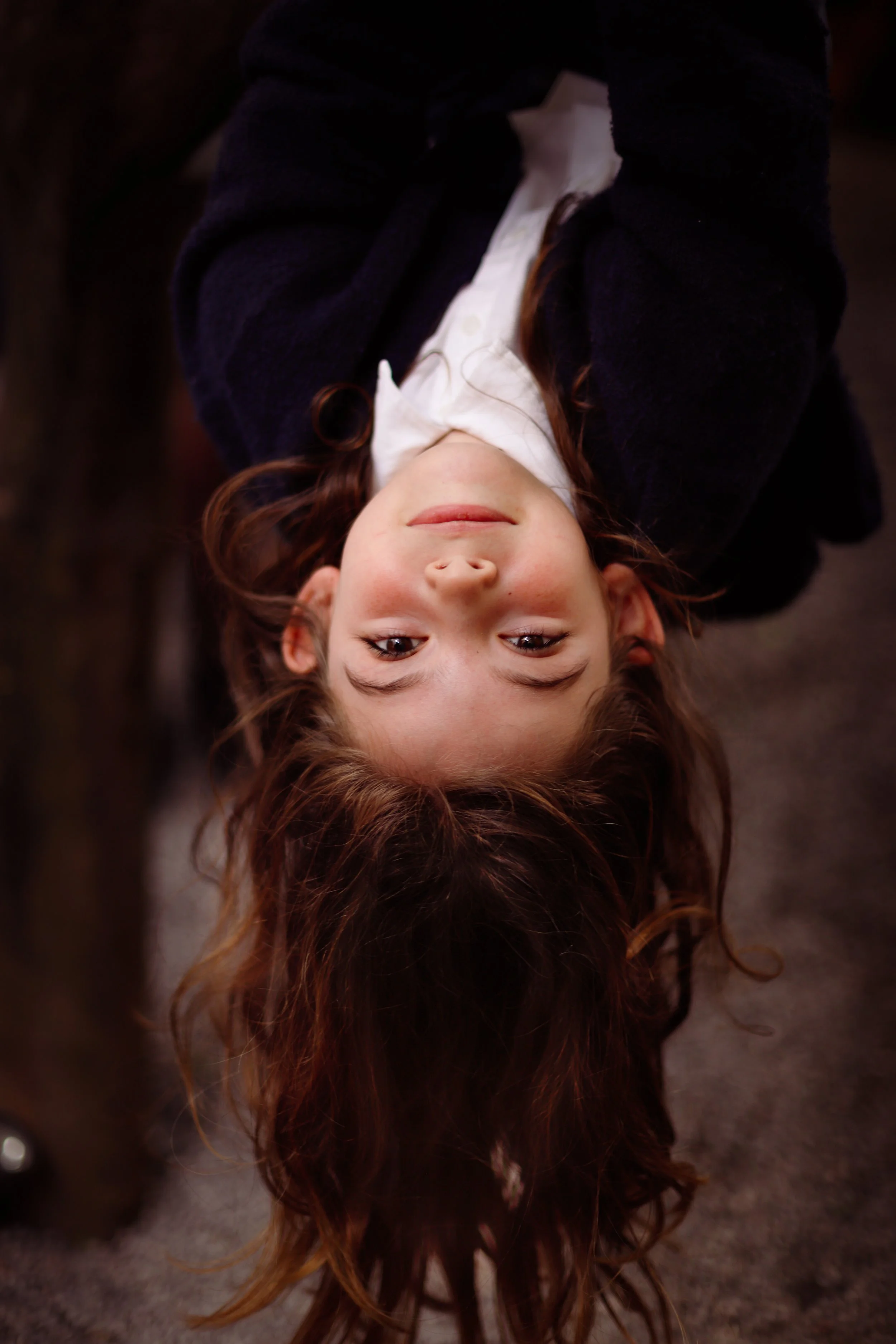 A young girl with curly brown hair lying upside down on the ground, smiling softly at the camera, with her head on the dirt and her eyes looking directly at the camera.
