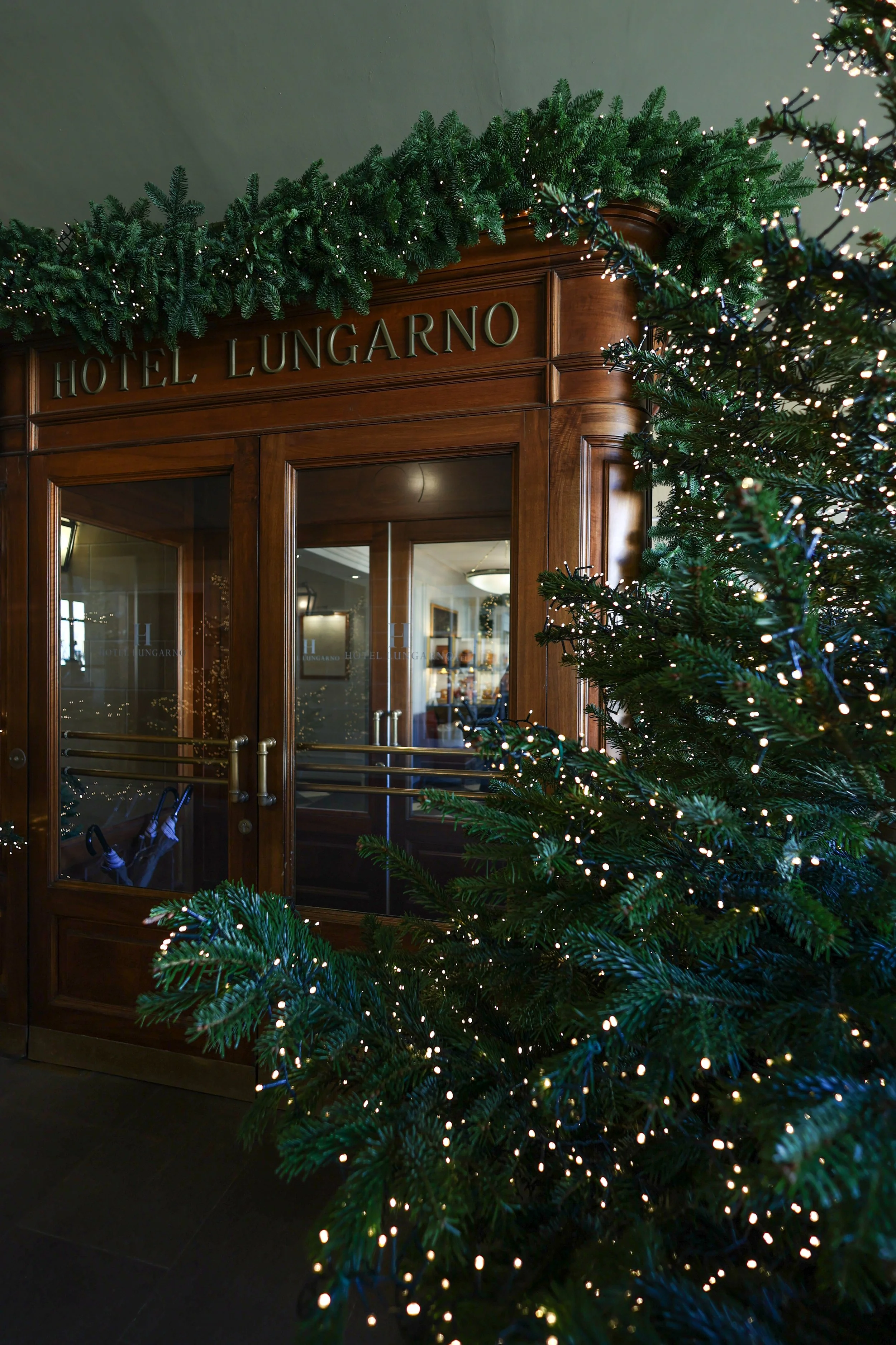 Decorative Christmas garland hanging above a wooden entrance door with glass panels, with a Christmas tree decorated with lights in the foreground at Lungarno Hotel in Florence