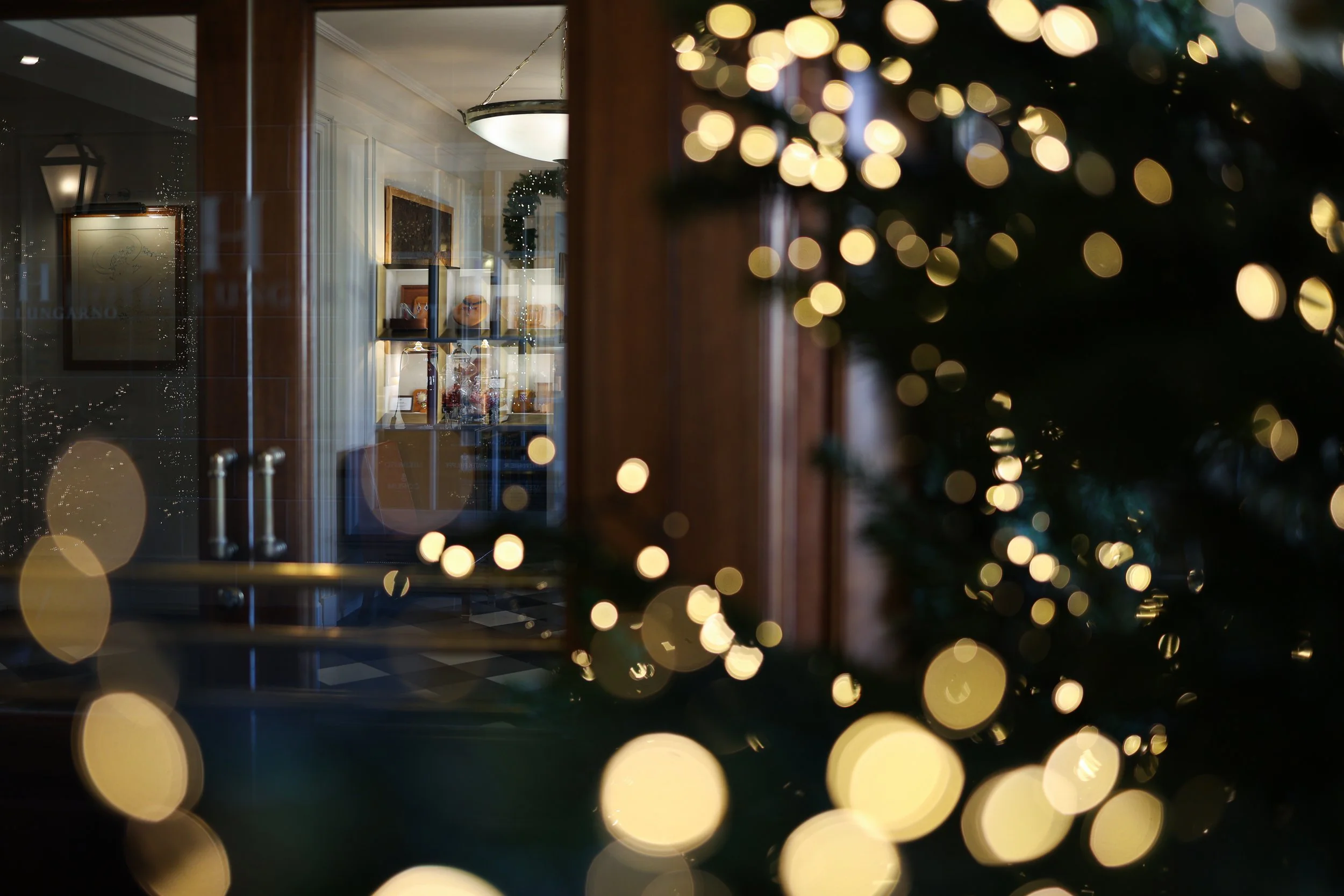 A blurred view of a room seen through a window adorned with Christmas lights, reflecting holiday decorations, a framed artwork, and a hanging light fixture inside, creating a festive atmosphere.