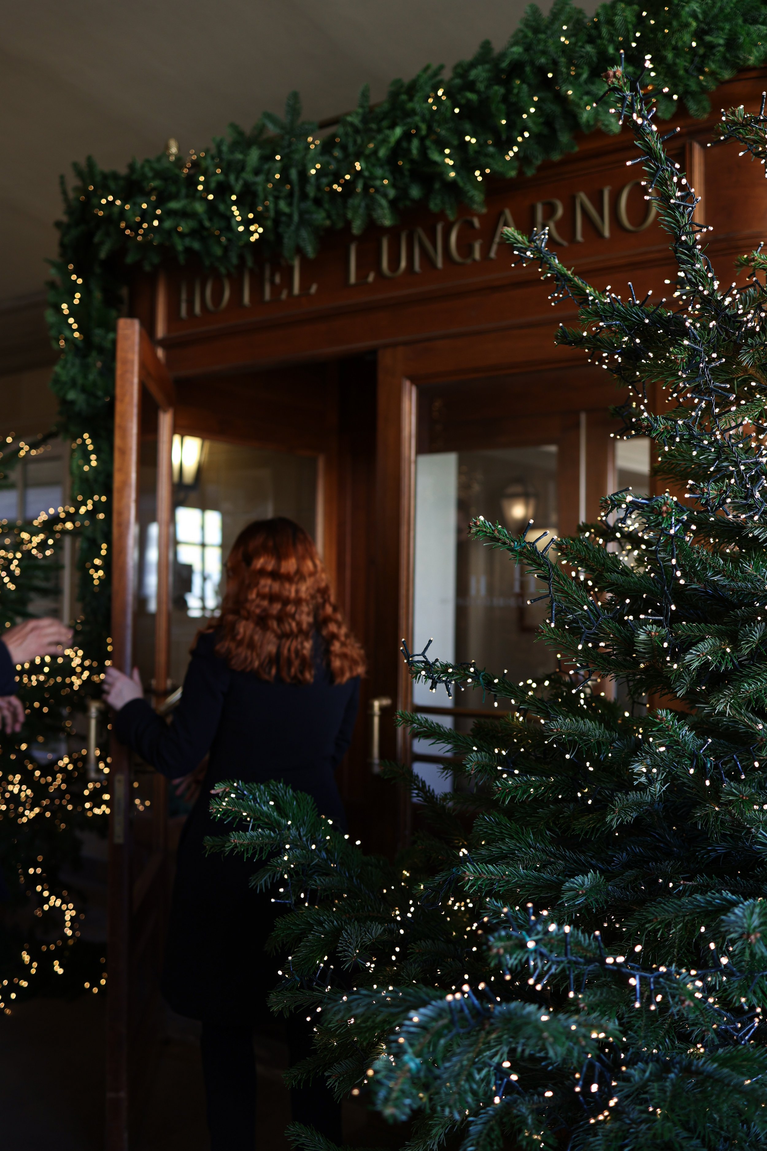 Christmas decorations with a decorated Christmas tree, green garland with string lights, and people inside Lungarno hotel lobby preparing for a holiday event.
