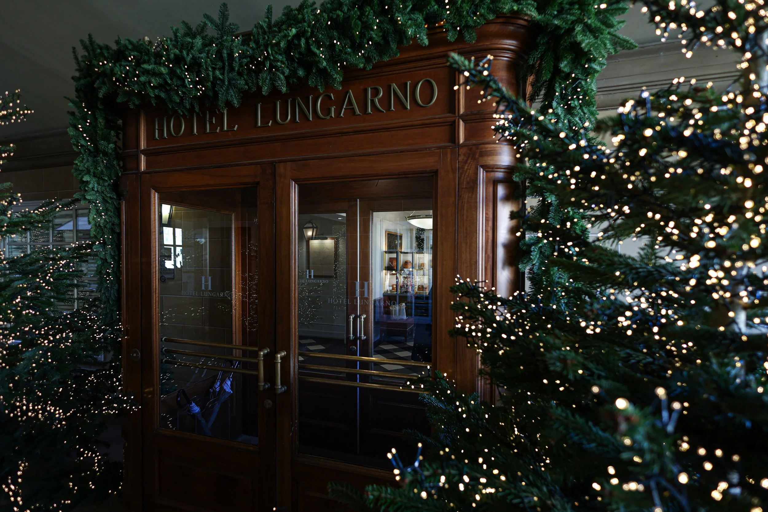 The image shows the entrance of Hotel Lungarno decorated for Christmas with green garlands and string lights. The hotel has a wooden framed glass door, and inside, there are framed pictures and a bright interior.