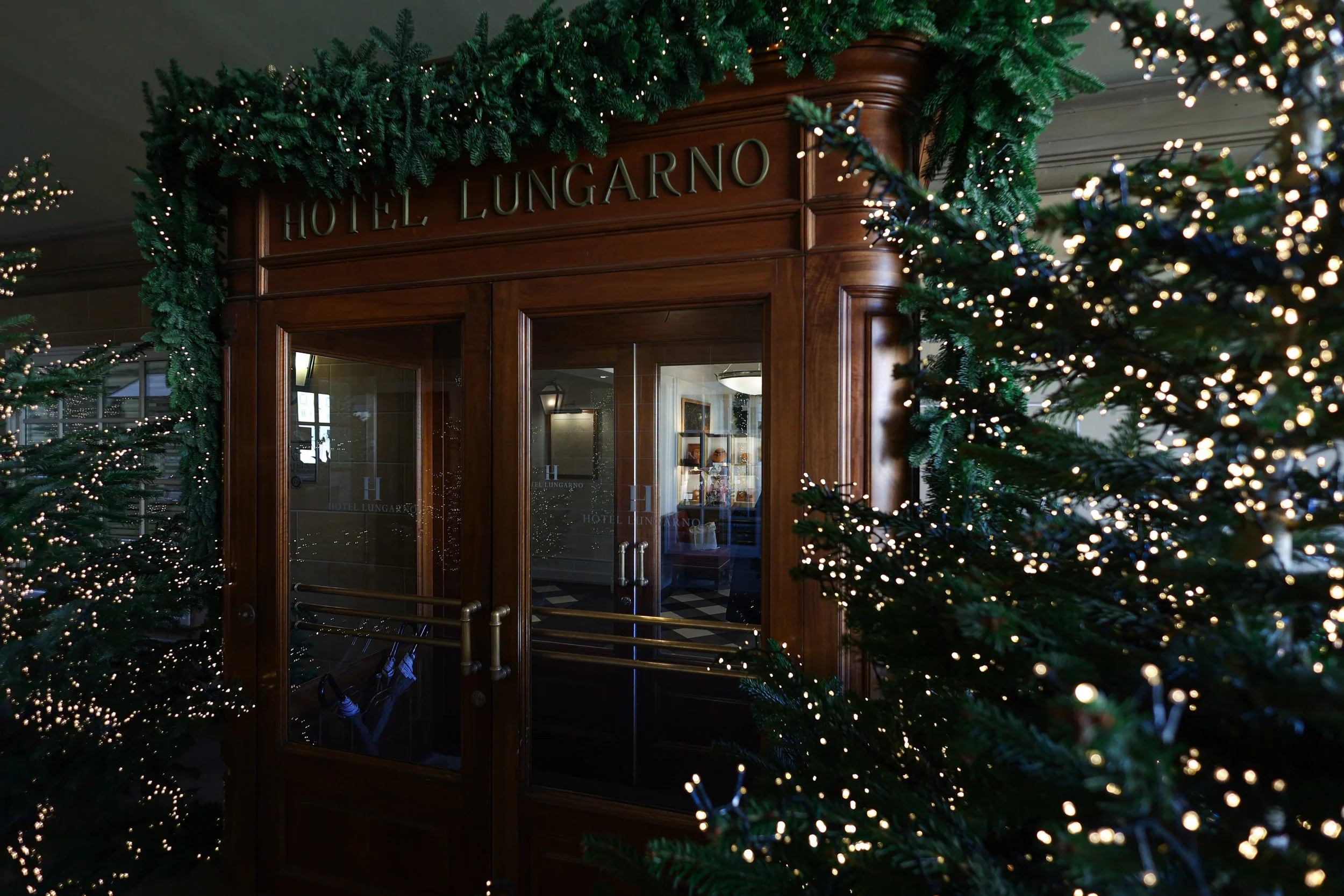 Decorated christmas trees with string lights flanking the entrance of Hotel Lungarno in Florence, which has a wooden door and sign.