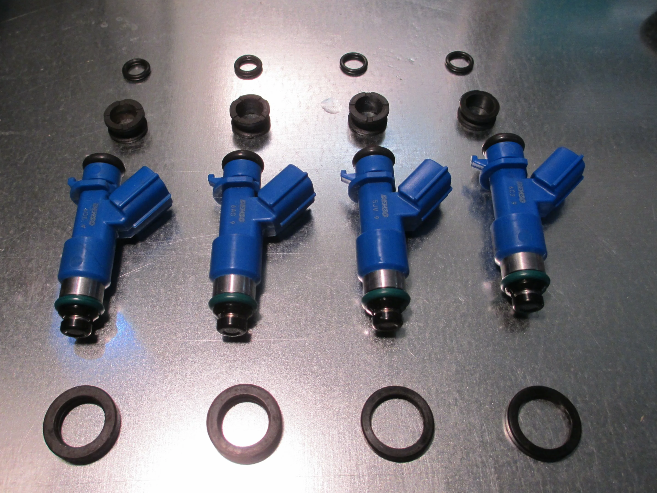 Blue fuel injectors with black rubber O-rings and metal tips are arranged on a metallic surface, with additional black rubber rings placed underneath them.