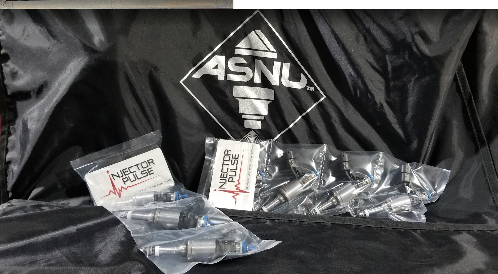 Multiple automotive fuel injectors in plastic bags, branded as 'Injector Pulse', resting on a black surface with a backdrop featuring a logo of a spark plug and the text 'ASNU'.