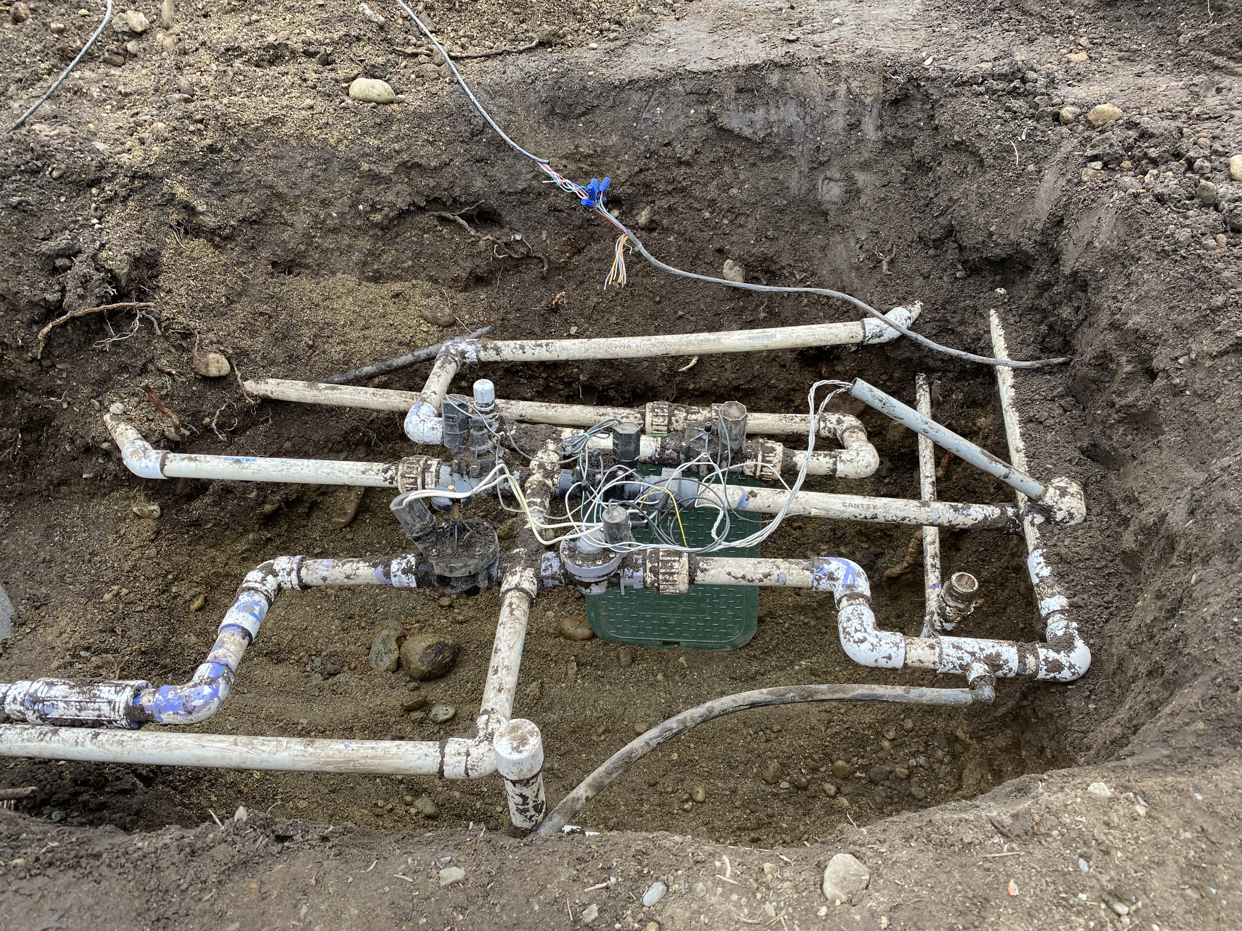 An open excavation site showing a network of exposed pipes with some electrical wiring attached, surrounded by dirt and small rocks.