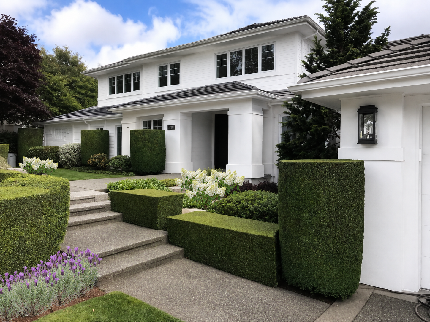 Front yard of a white house with landscaped garden, manicured bushes, and concrete steps leading to the entrance.