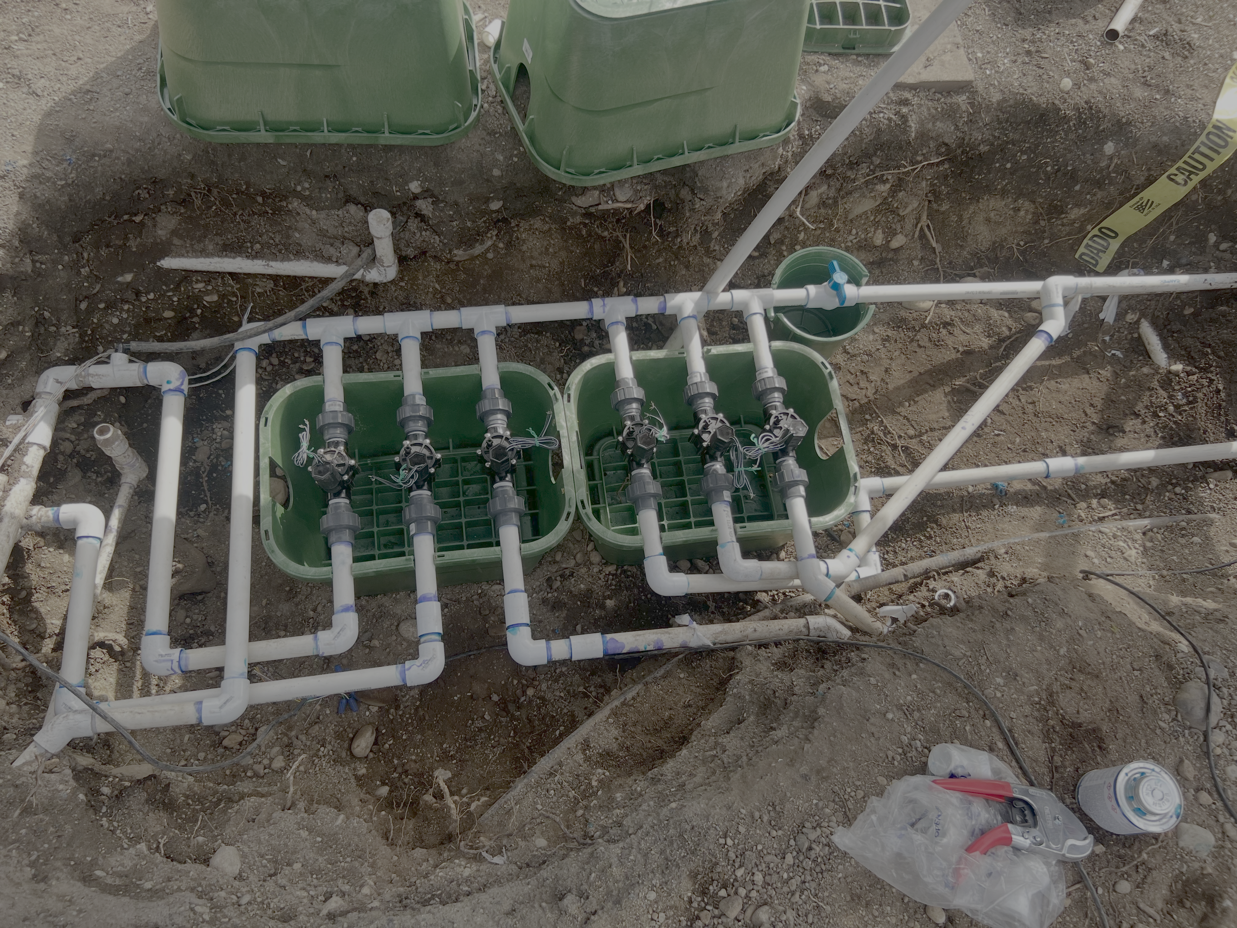 View of irrigation system with white pipes, green containers, and connected black valves on a dirt ground, possibly at a construction site.