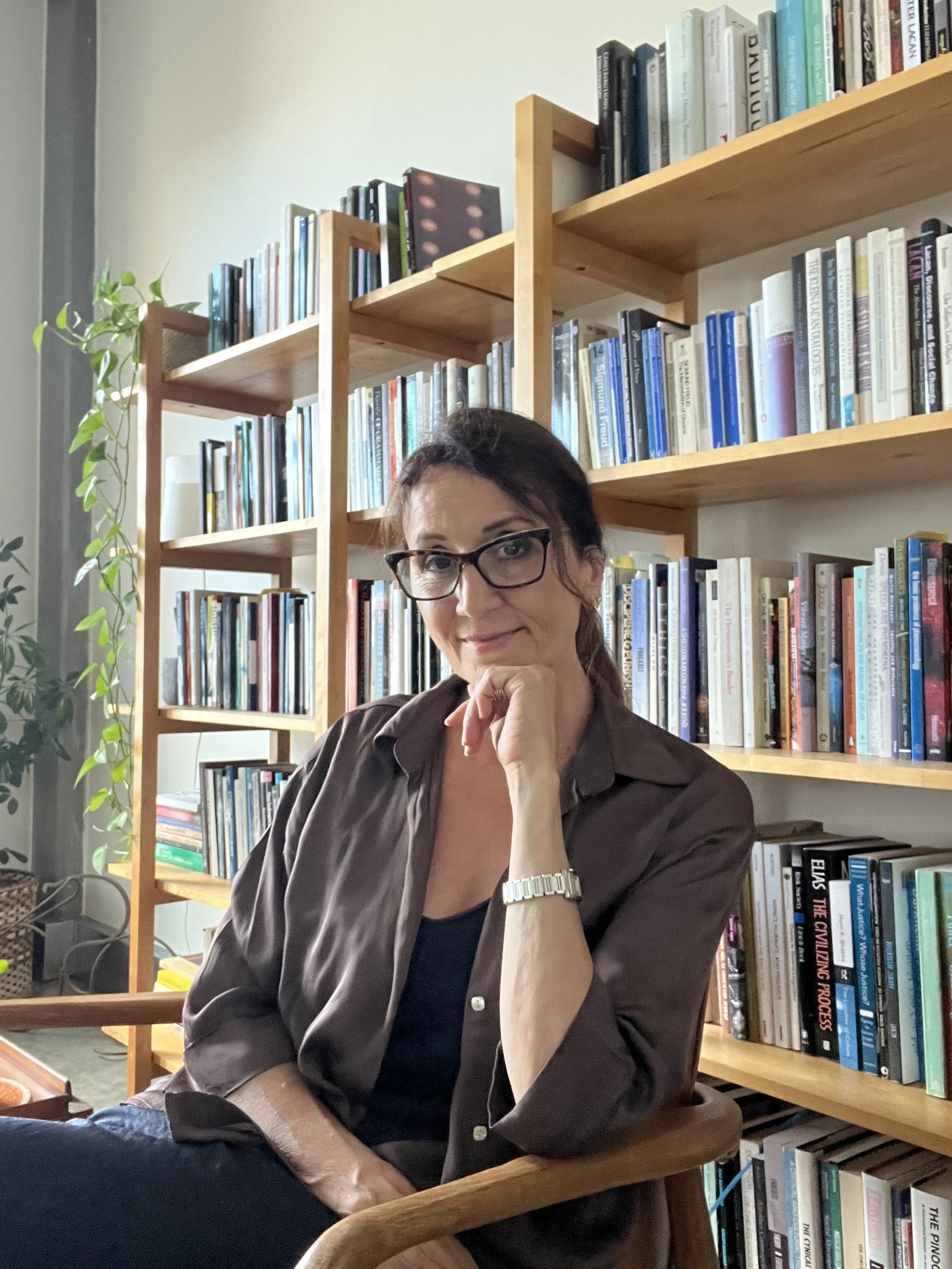 A woman with glasses sits in front of a bookshelf filled with books, resting her chin on her hand, with a plant on the left and a wooden table in the foreground.