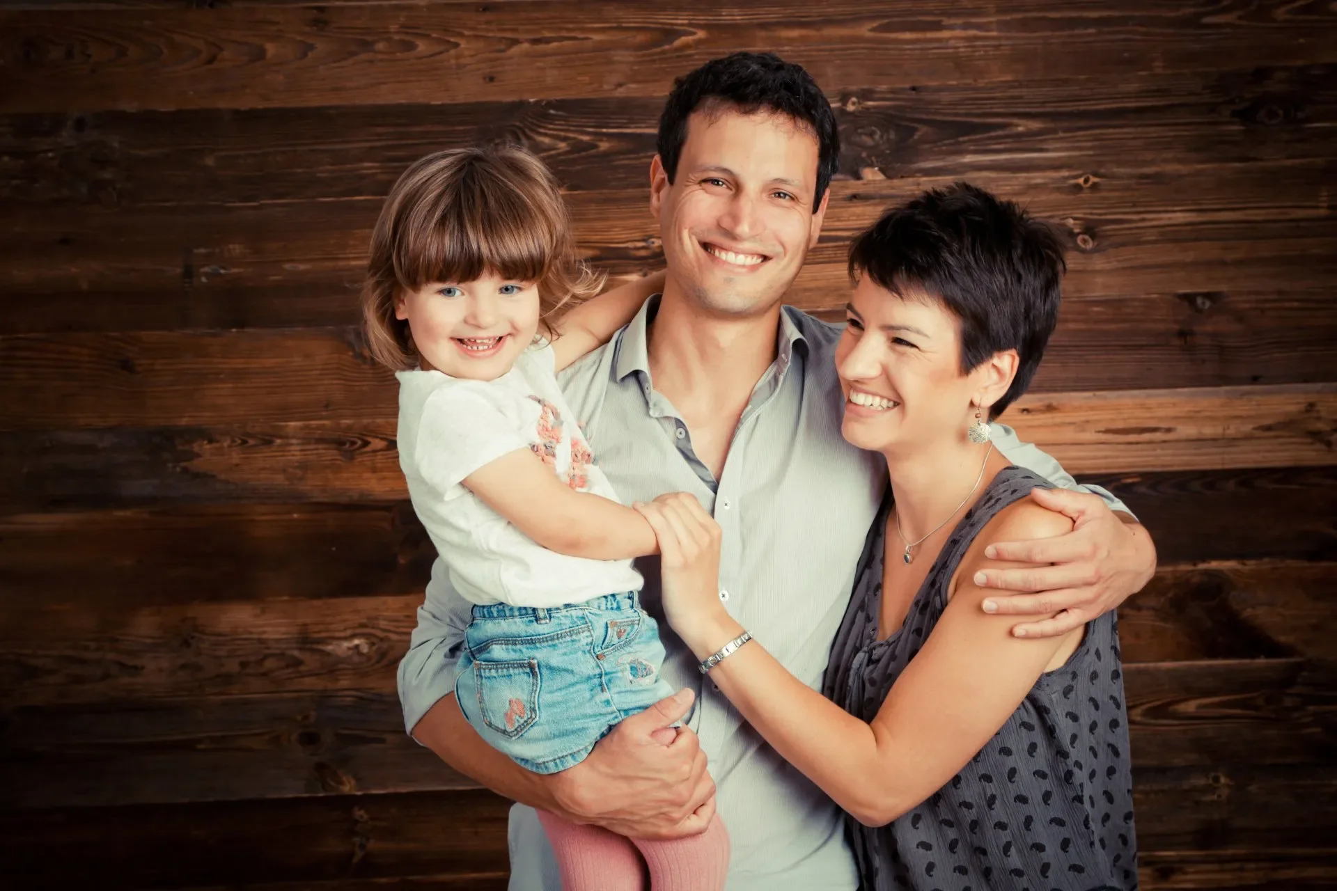 Portrait Aufnahme einer Familie bestehend aus einem Mann, einer Frau und einem Mädchen, die lachend vor einer Holzwand stehen und sich umarmen.