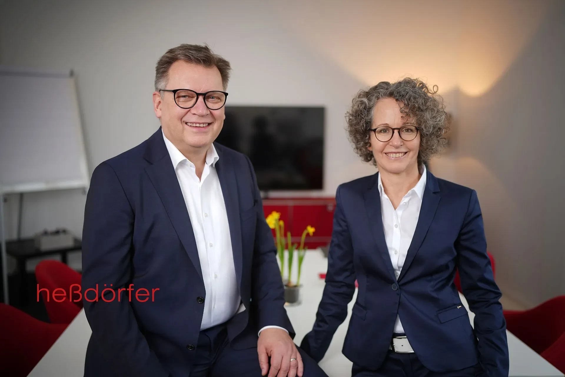 Ein Mann mit Brille und dunklem Anzug steht neben einer Frau mit Brille und dunklem Anzug, beide lächeln in die Kamera, im Hintergrund ein Fernseher und eine rote Wand. 