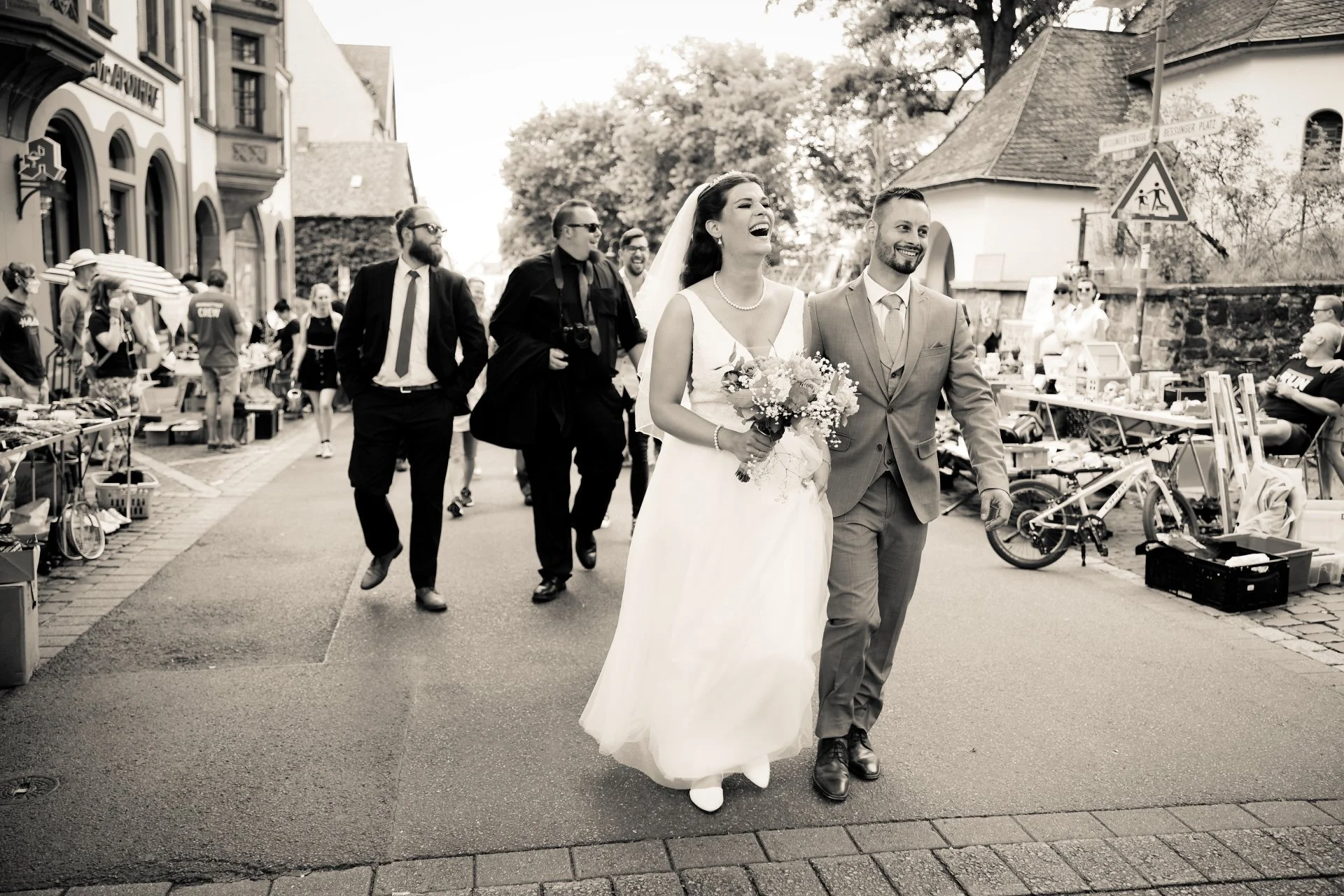 Ein glückliches Brautpaar, die Braut in einem weißen Kleid mit Blumengesteck und der Bräutigam in einem grauen Anzug, geht lachend die Straße entlang, während Gäste und Verkäufer im Hintergrund bei einem Straßenmarkt zu sehen sind.