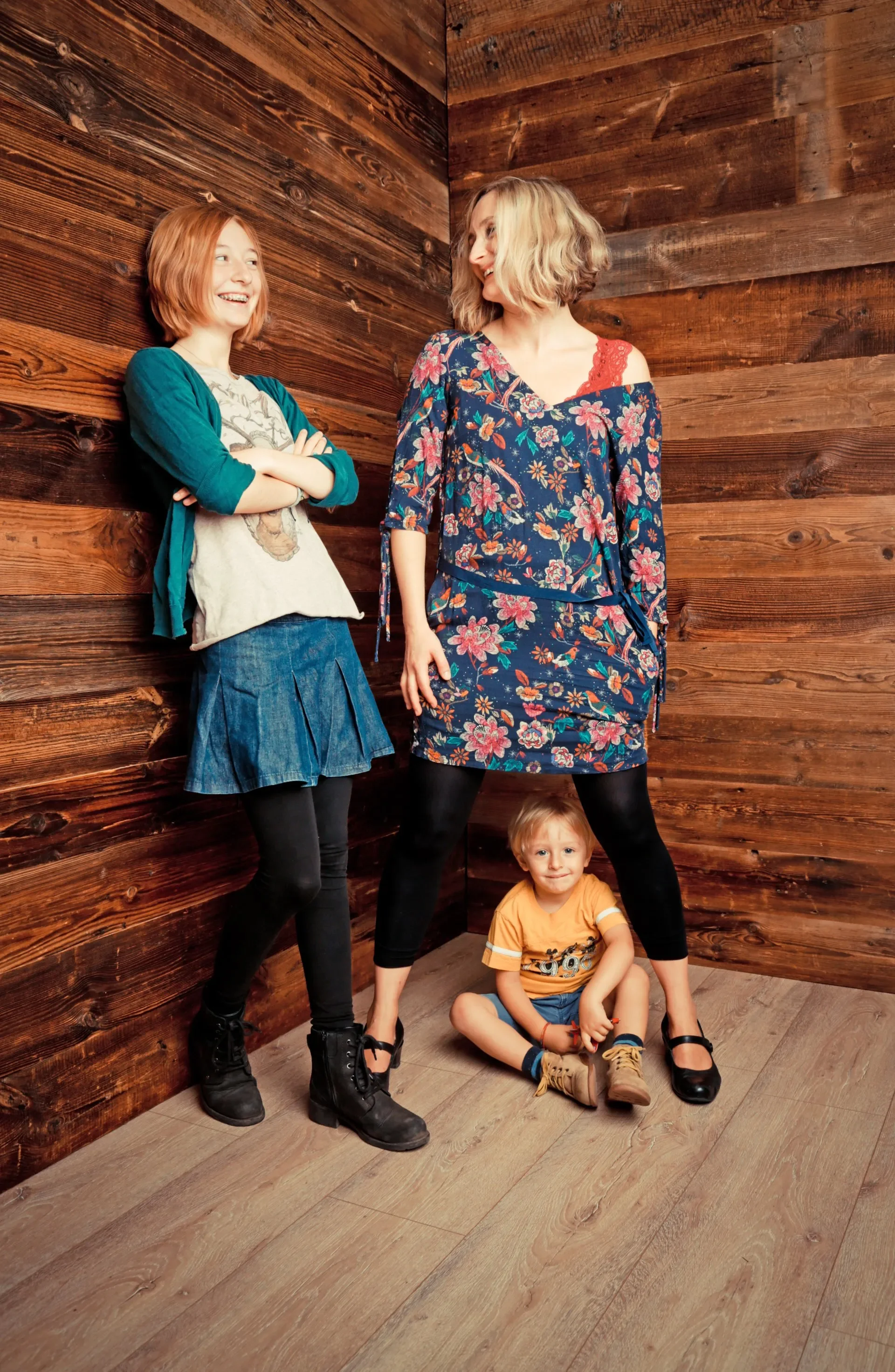 Familien Portrait Fotoshooting - Kinder posieren vor einer Holzvertäfelung. Zwei Mädchen stehen, ein Junge sitzt am Boden. Alle lachen und wirken fröhlich.