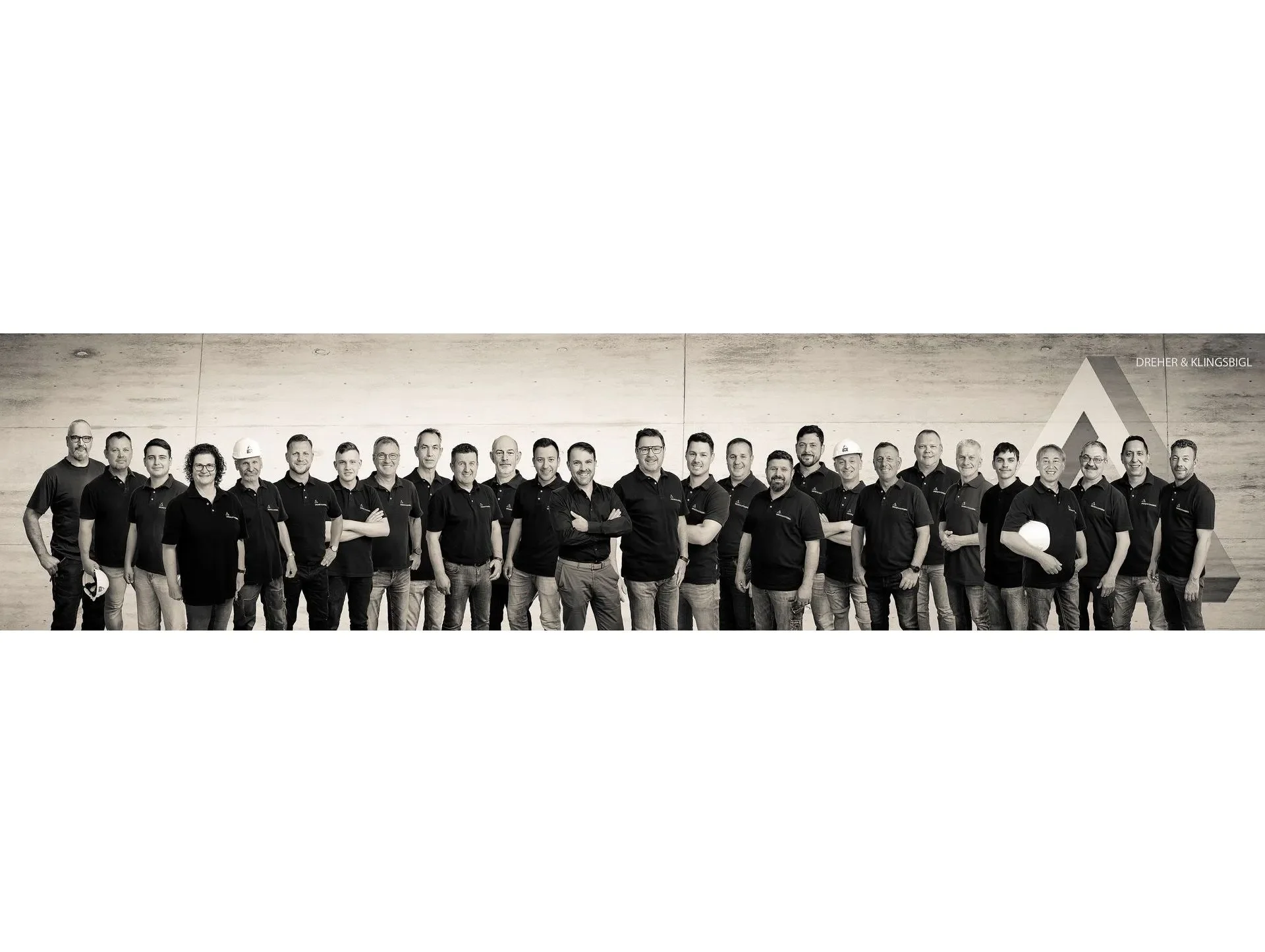 Gruppe von Menschen trägt schwarze Polohemden, steht nebeneinander vor einer Wand, einige halten Helme, alle lächeln, im Hintergrund eine Stehleuchte und eine Wand aus Holzplatten.