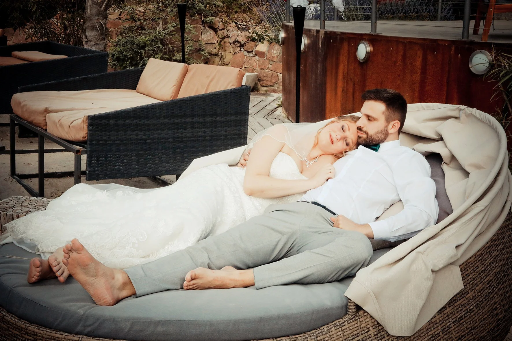 Ein frisch verheiratetes Paar liegt entspannt auf einem kuscheligen Liegestuhl im Garten, sie trägt ein Hochzeitskleid, er ein Hemd und Anzughose, sie schauen sich liebevoll an.