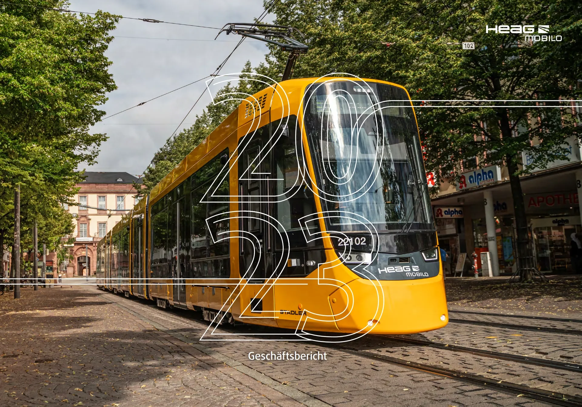 Ein gelber Straßenbahnzug fährt auf den Schienen entlang einer Baumallee in Darmstadt. Im Hintergrund befinden sich Geschäfte und Gebäude. Über der Bahn sind elektrische Kabel zu sehen.