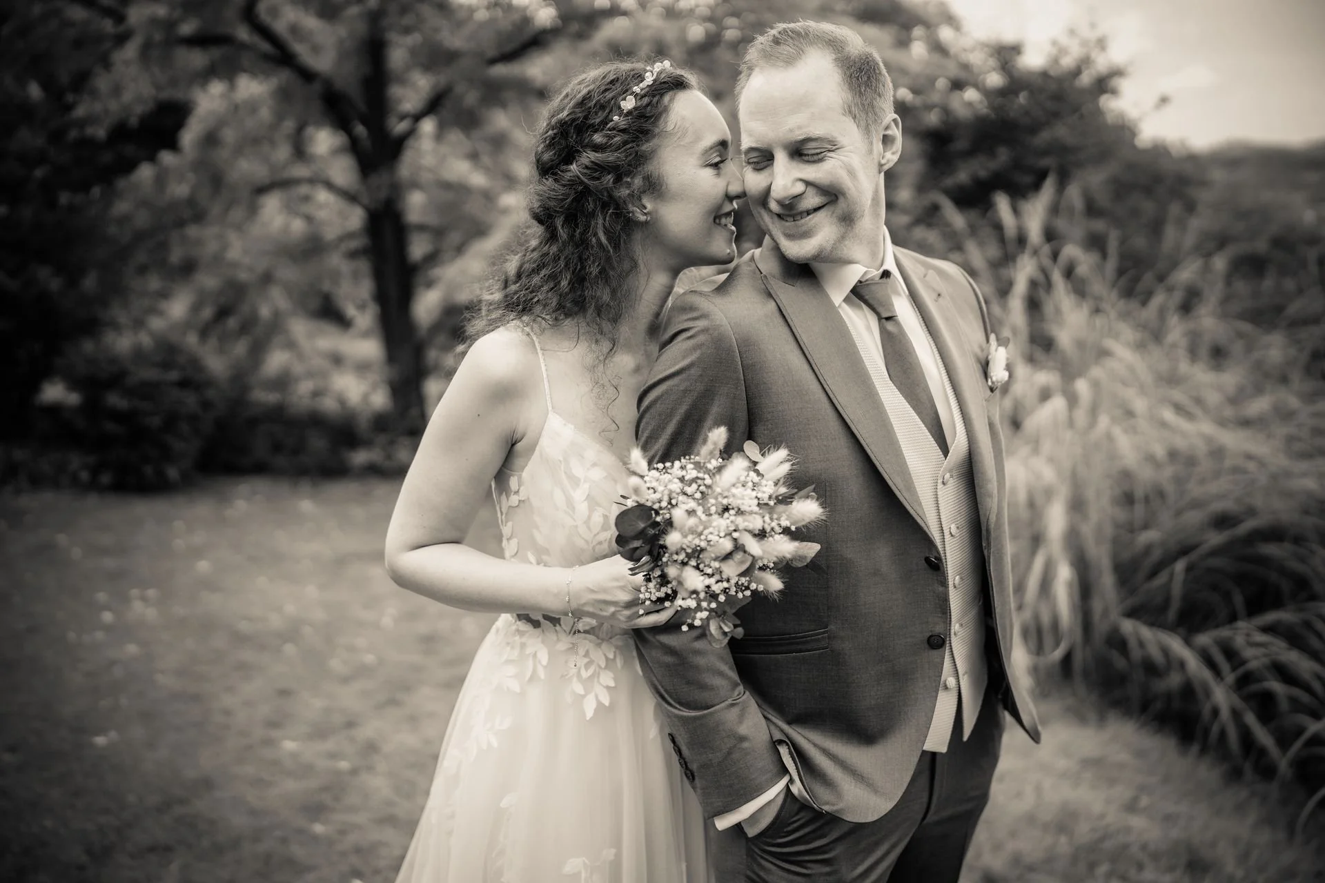 Hochzeitsfoto von Braut und Bräutigam beim Hochzeitspaar in einem Park, schwarz-weiß Fotografie, die Braut hält einen Blumenstrauß, beide lächeln sich an.