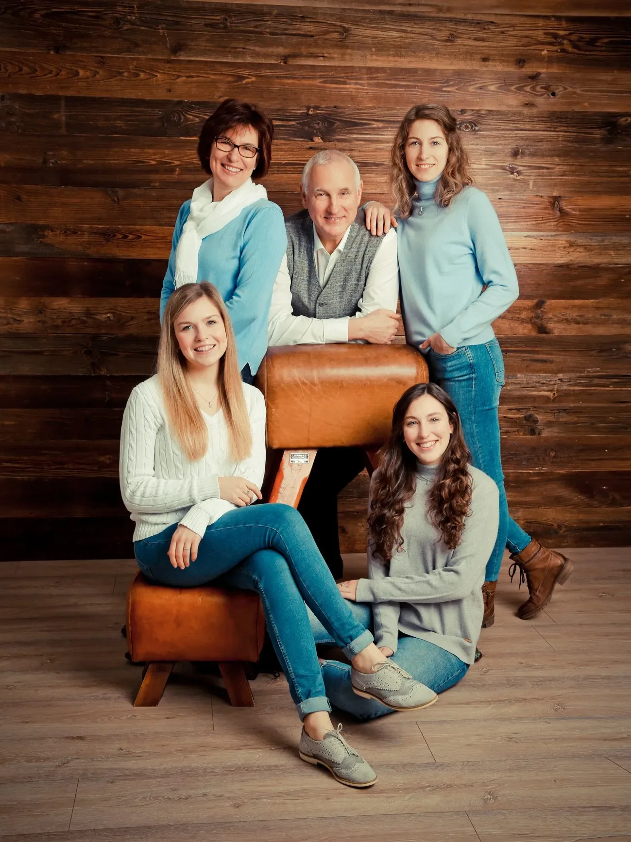 Familien Portrait - Familie mit vier jungen Erwachsenen vor einer Holztapete. Zwei Frauen sitzen auf einem Lederhocker, eine Frau steht hinter einem Mann, eine weitere Frau sitzt auf dem Boden.
