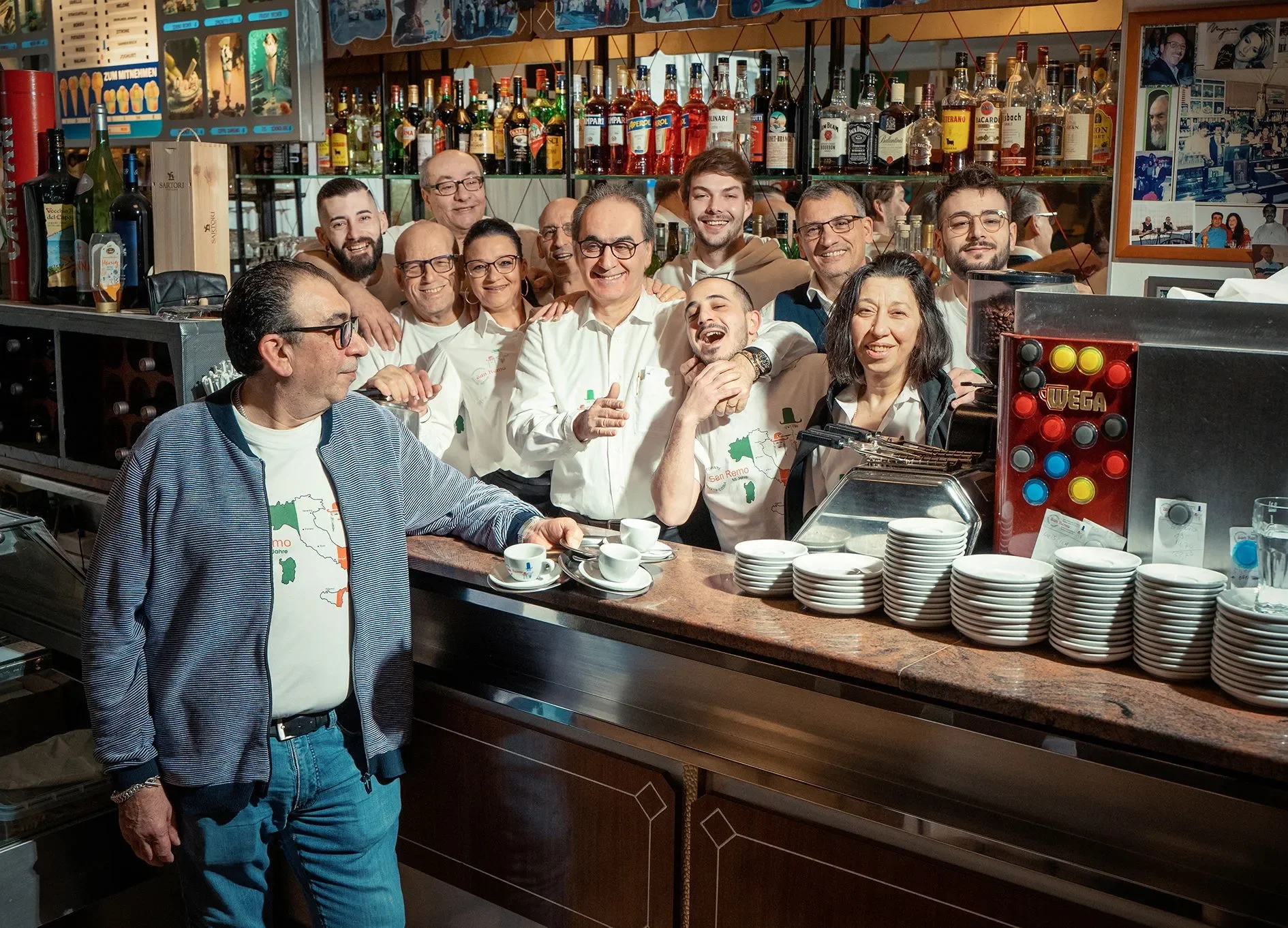 Eine Gruppe von Menschen, die im Barbereich eines Restaurants oder Cafés stehen, lachen und posieren für das Foto. Der Hintergrund zeigt eine Bar mit zahlreichen Spirituosenflaschen und Dekorationen. Im Vordergrund steht ein Mann in Jeans und einer J