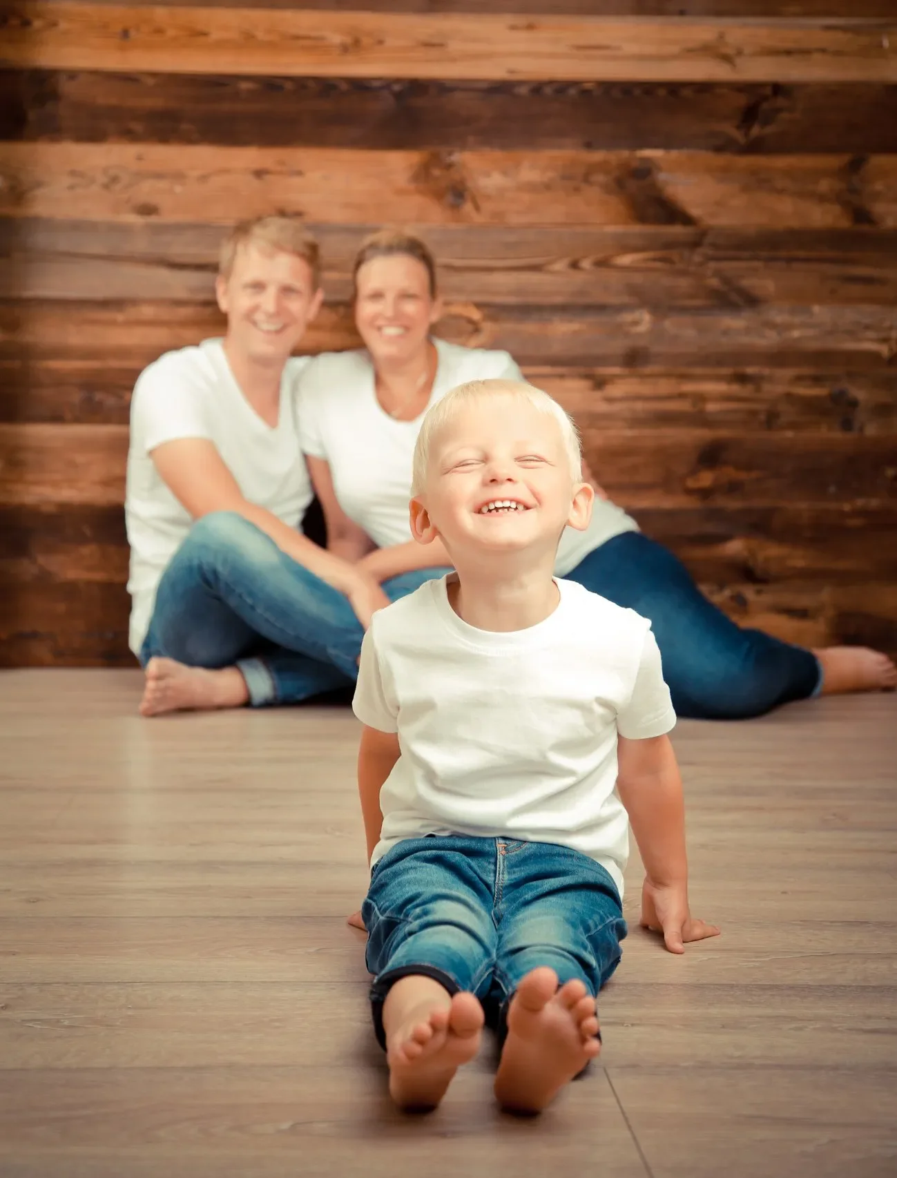 Familien Portrait Ein lachendes Kind sitzt auf dem Boden mit erhobenem Oberkörper, im Hintergrund ein Paar, das auf dem Boden sitzt. Die Szene ist in einem Raum mit Holzvertäfelung hinter dem Paar.