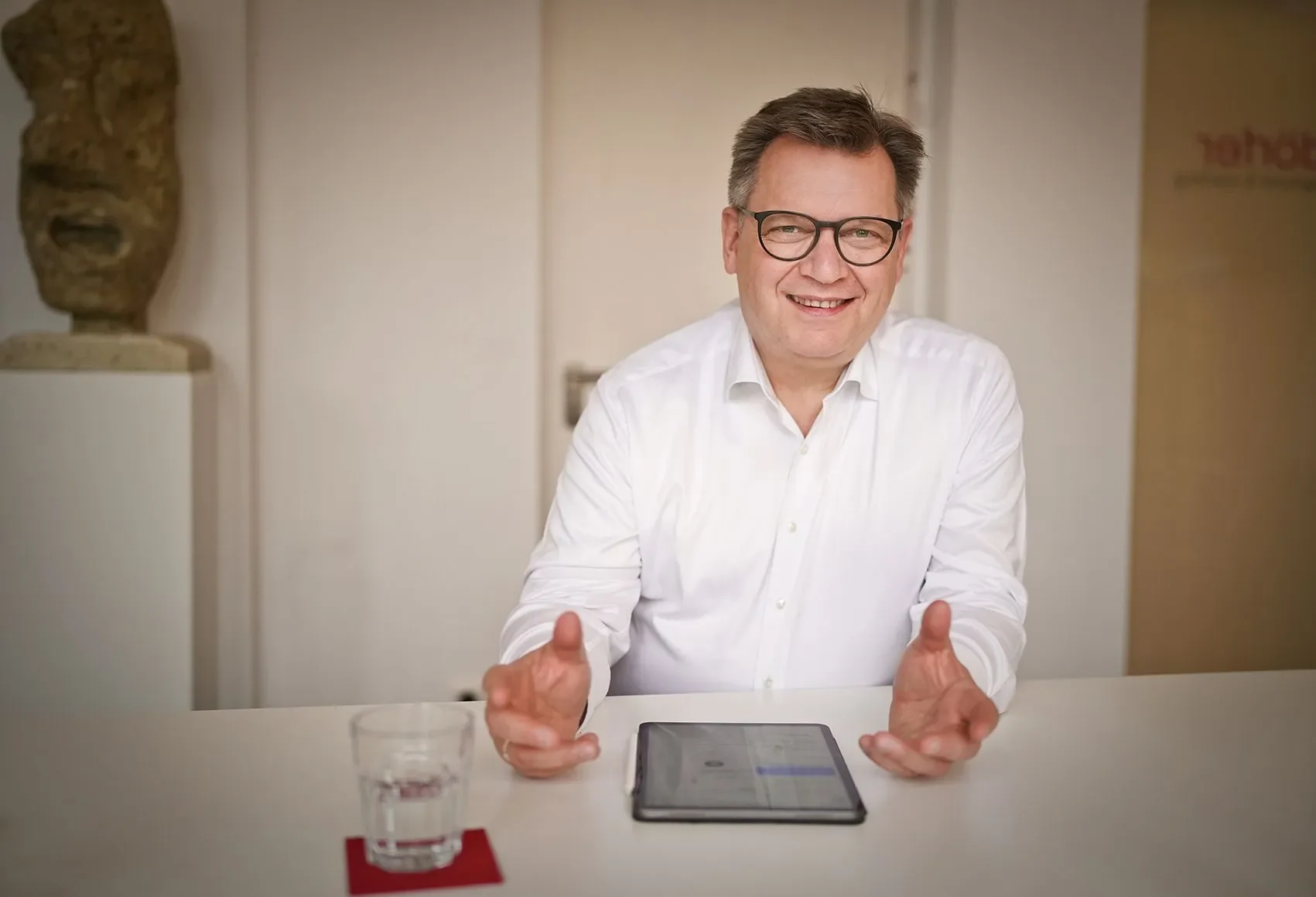 Ein Mann sitzt an einem Tisch, lächelt und gestikuliert mit den Händen, vor ihm liegt ein Tablet. Auf dem Tisch steht ein Glas Wasser auf einem roten Untersetzer.