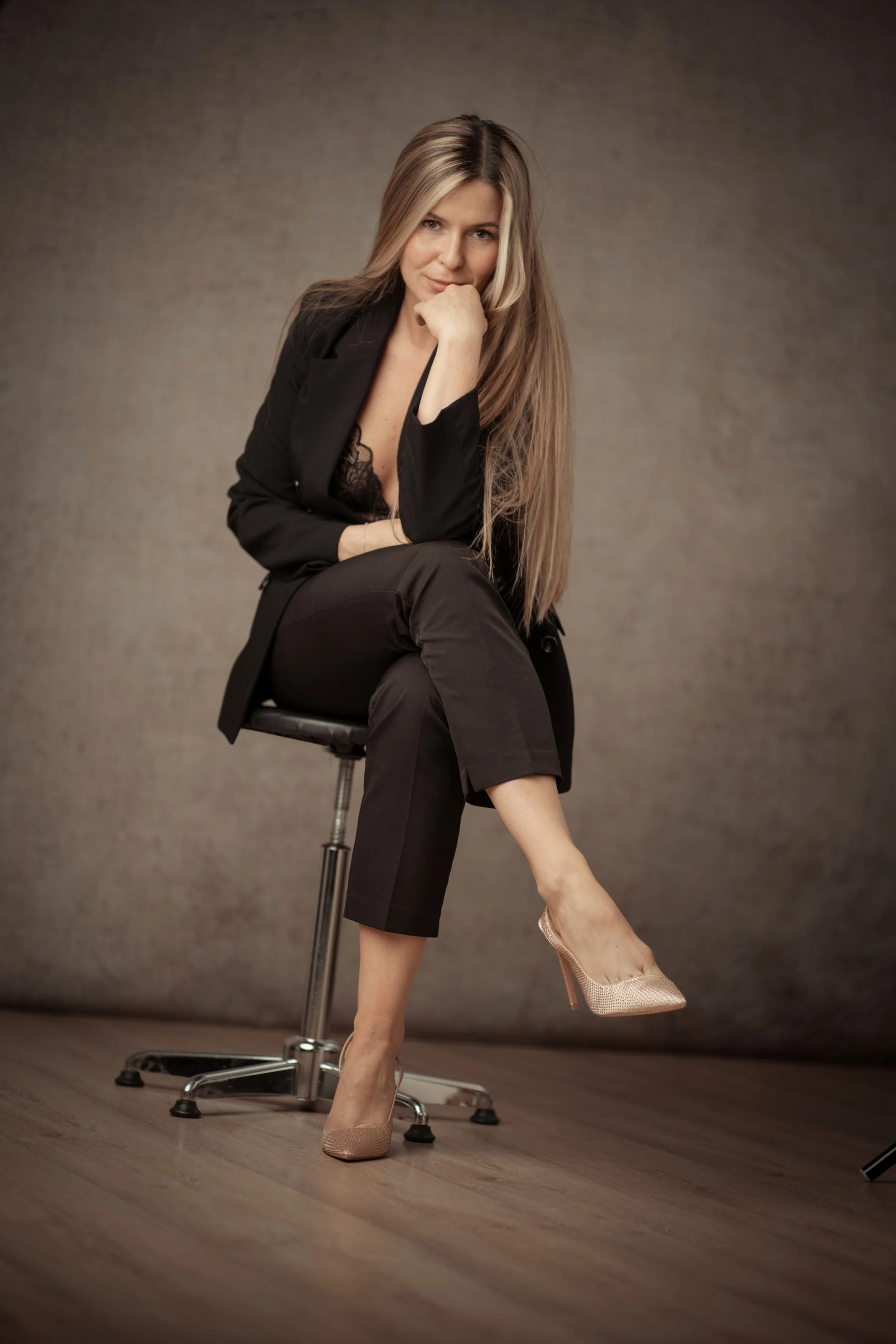 Frau in schwarzer Kleidung sitzt auf einem Stuhl, hebt ein Bein in einem wirksamen Posing, trägt high heels und blickt in die Kamera.